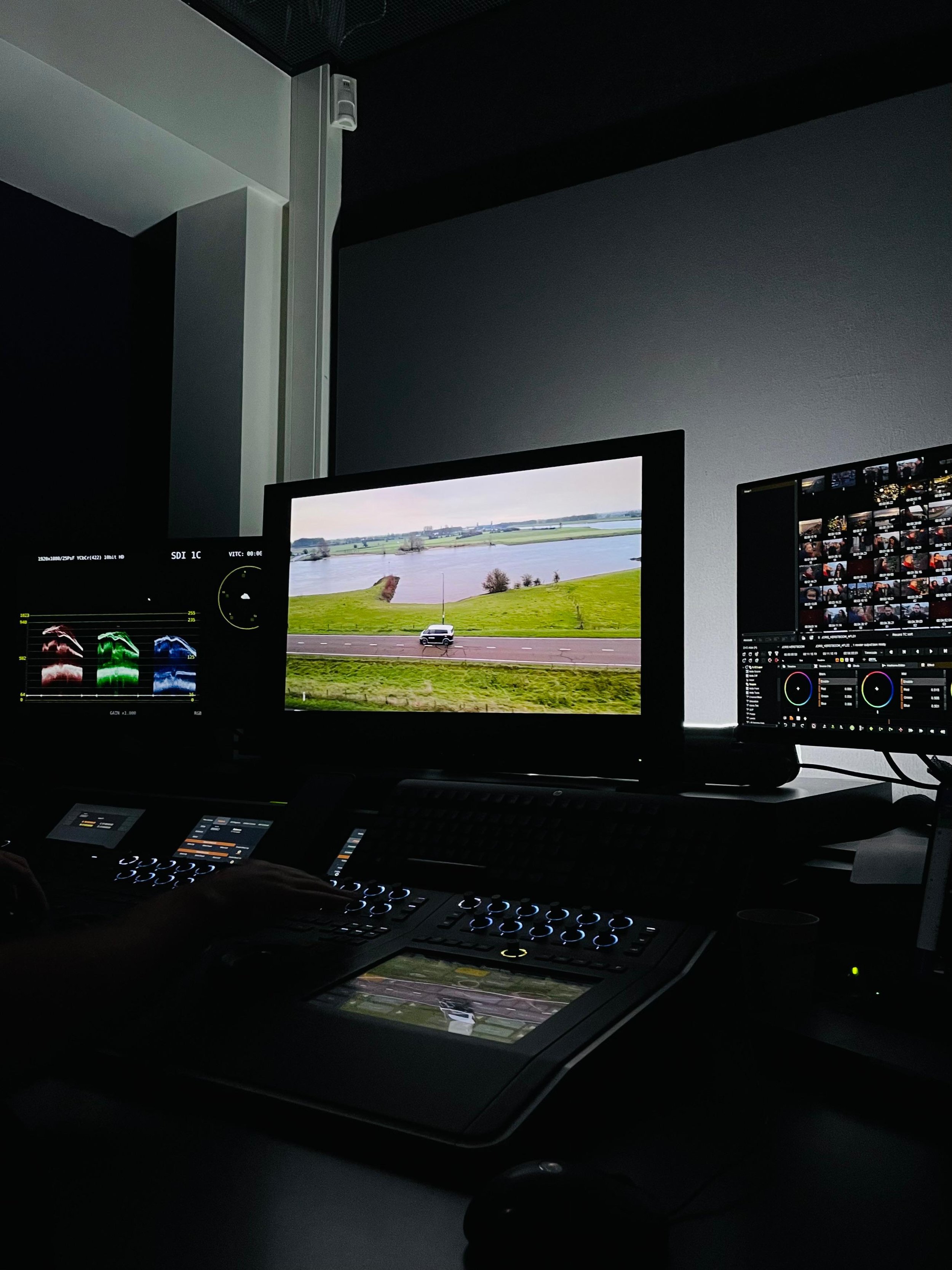 Een controlekamer met meerdere computerschermen, waarvan één scherm een landschap met water en groen toont, en andere schermen vuren foto’s en images van videobewerking en fotobewerking. Een persoon gebruikt een controlepaneel met knoppen en een touchscreen.