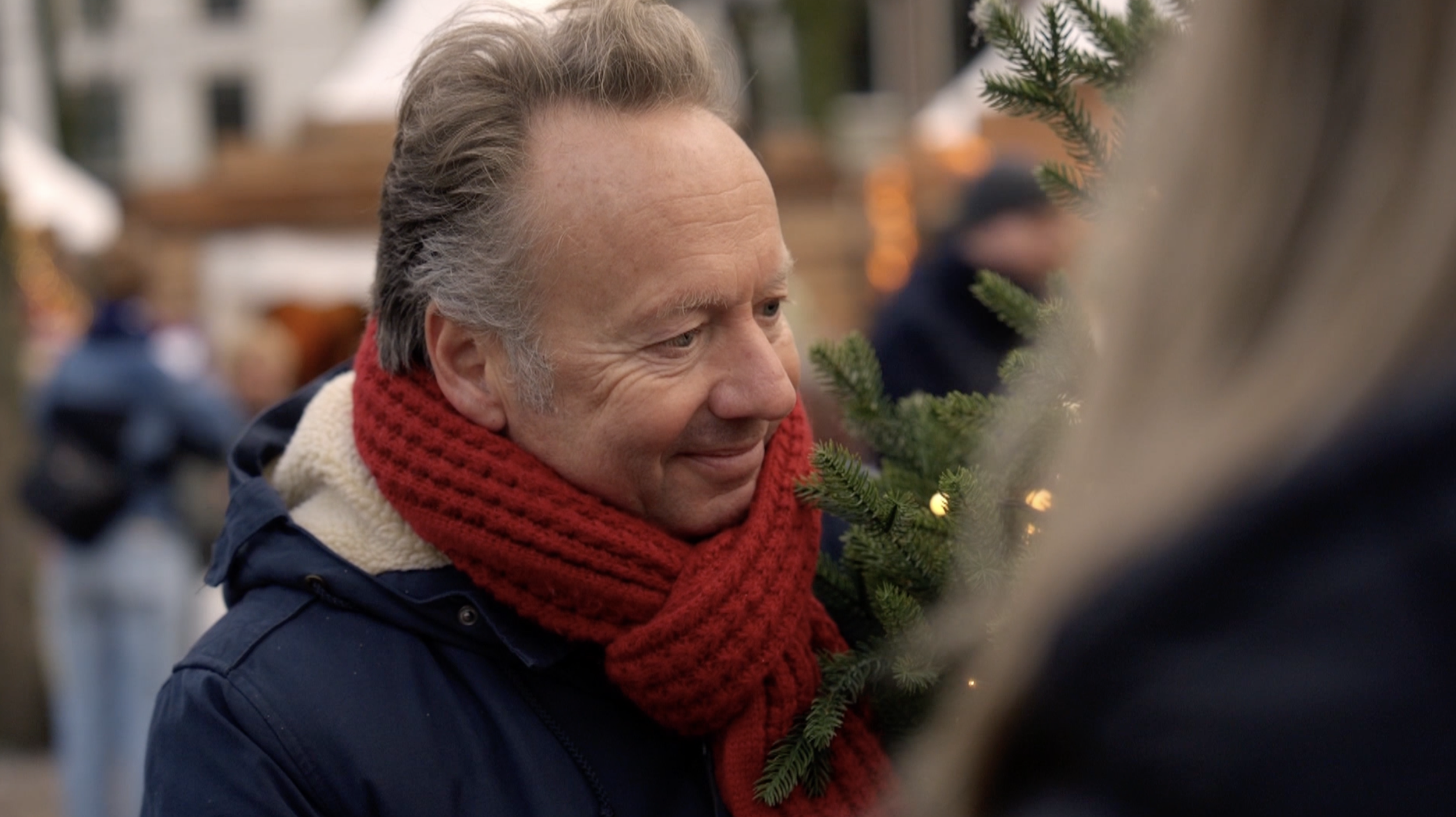 Man met grijs haar en een rode sjaal glimlacht terwijl hij naar een dennenboom kijkt in een kerstmarkt.