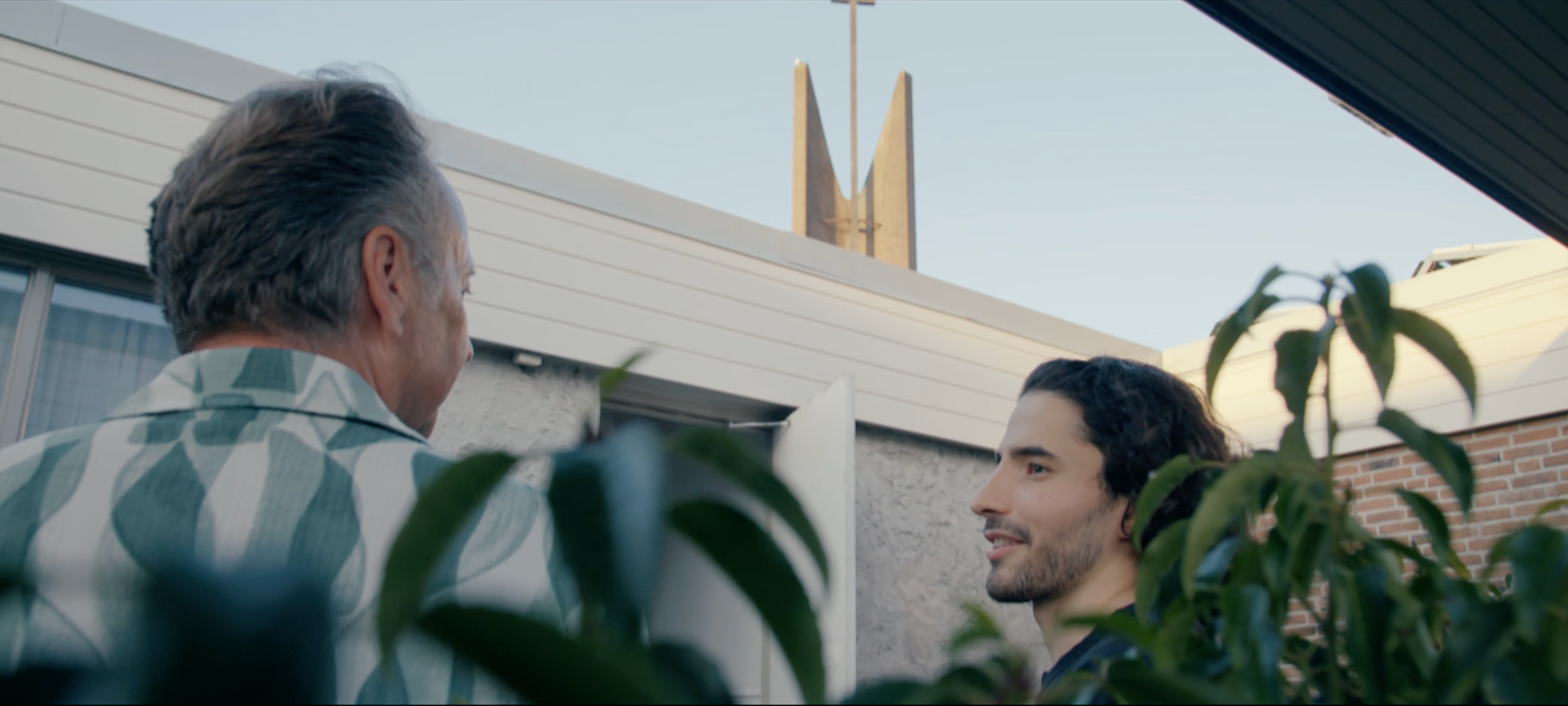 Twee mannen praten buiten, omgeven door groene planten, met huizen en een gebouw met een kerkklok op de achtergrond.