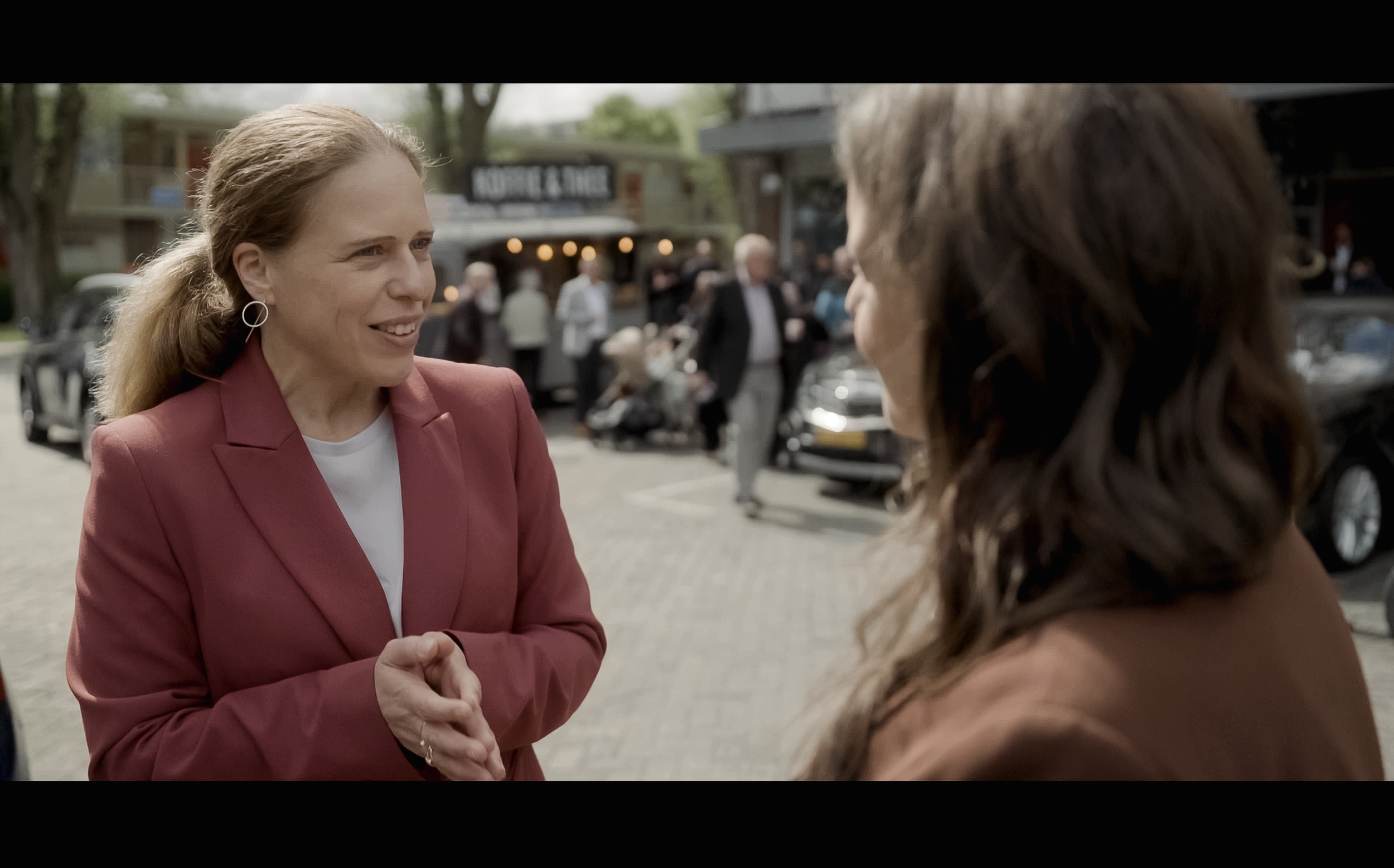 Twee vrouwen knikken elkaar de hand en glimlachen, buiten in een uitgaansgebied met mensen, auto's en winkels op de achtergrond. Burgemeester Carola Schouten