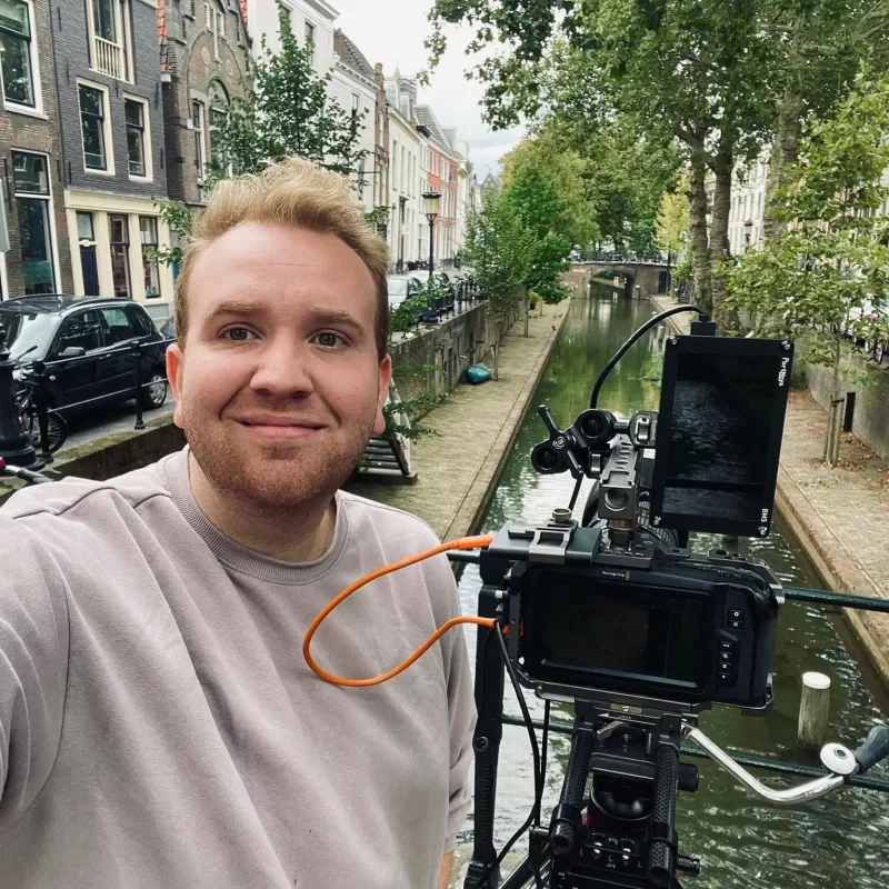 Een man neemt een selfie voor een grachten in een stad, met een camera op statief en veel bomen en oude gebouwen op de achtergrond.