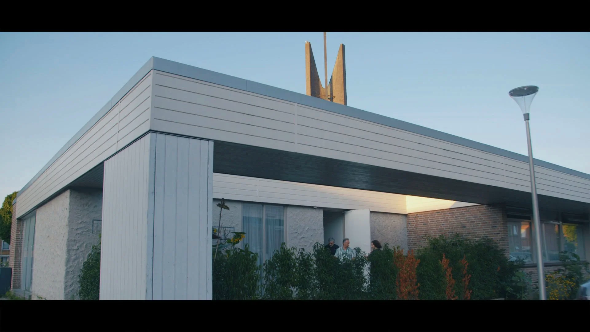 Modern wit en beton huis met veranda en tuin, drie mensen praten buiten, straatlantaarn, blauwe lucht.