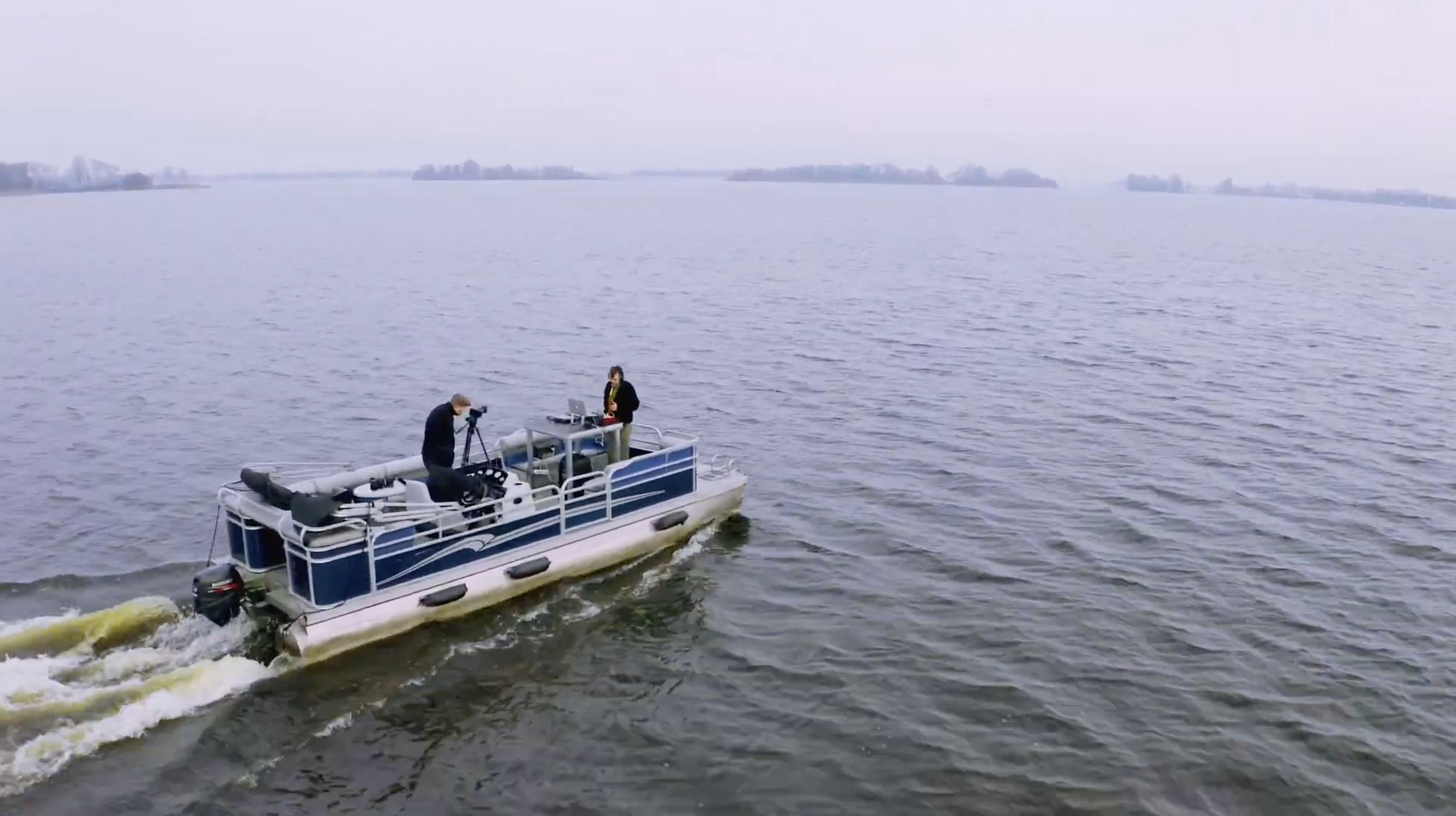 Twee mensen op een boot op een rustig meer met een laptop en camera, omgeven door een mistige en kalme omgeving.