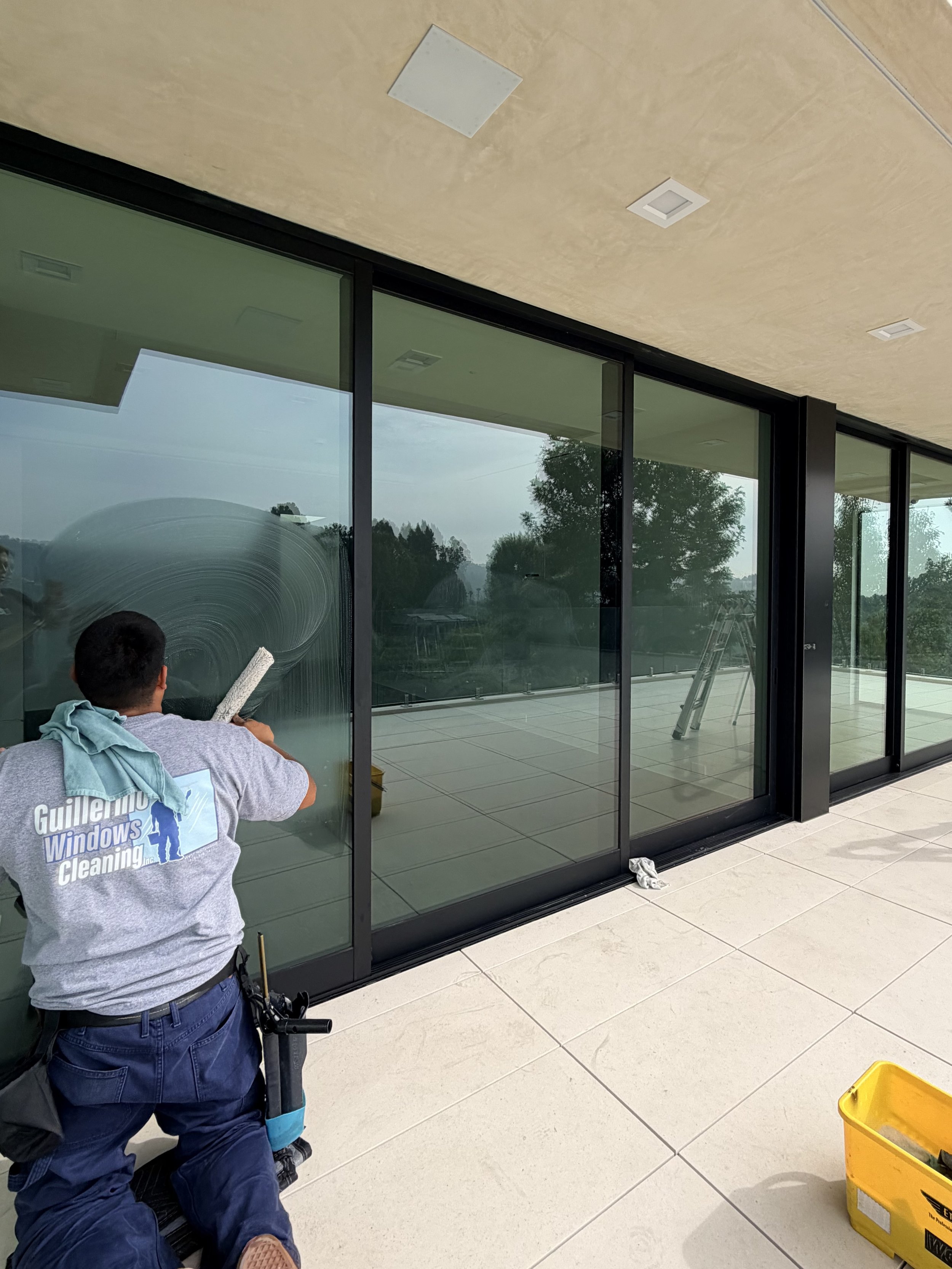 Window cleaning technician removing soap from large glass windows on a modern building with an outdoor patio and trees in the background.