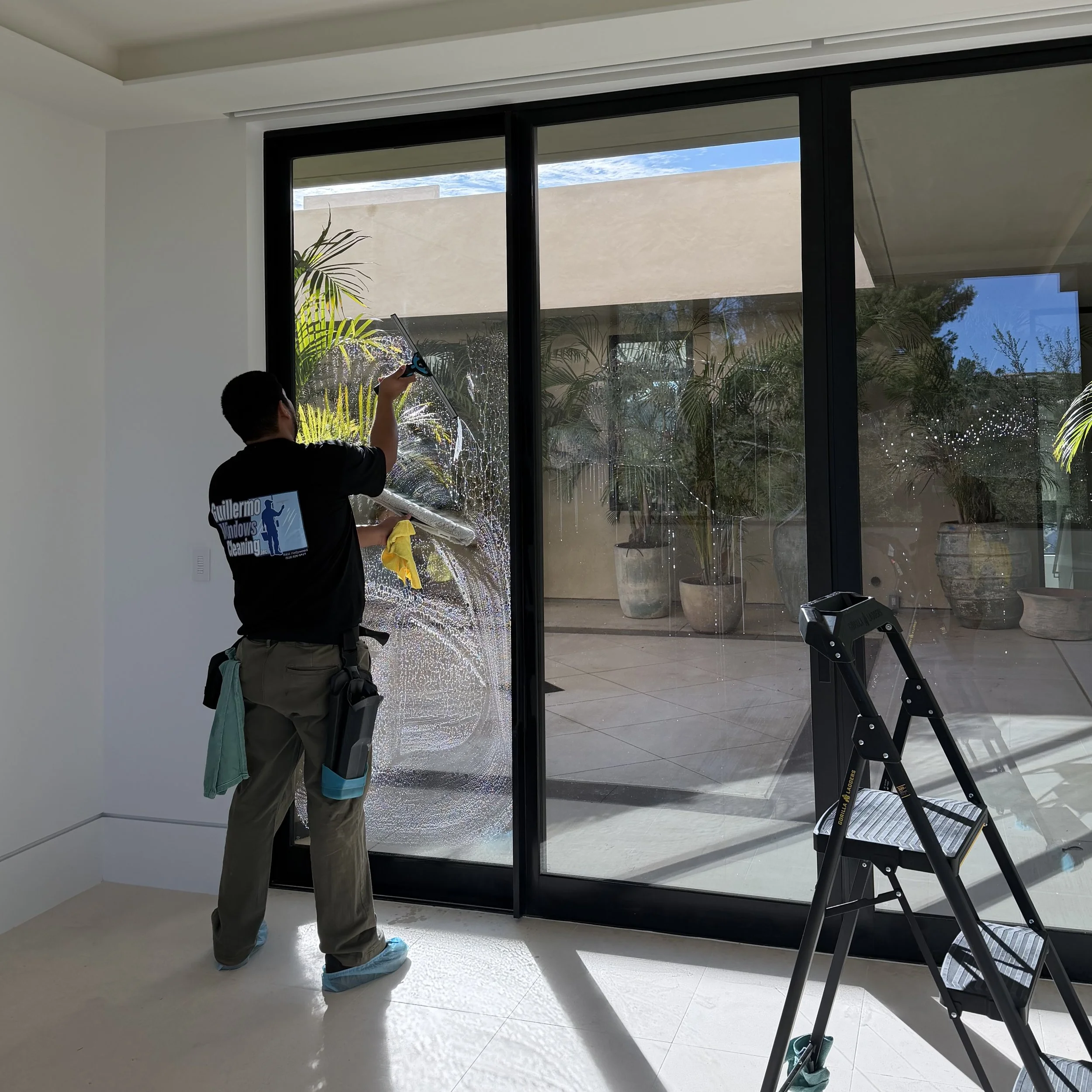 A cleaner is washing large glass sliding doors indoors using a squeegee, with sunlight streaming in.