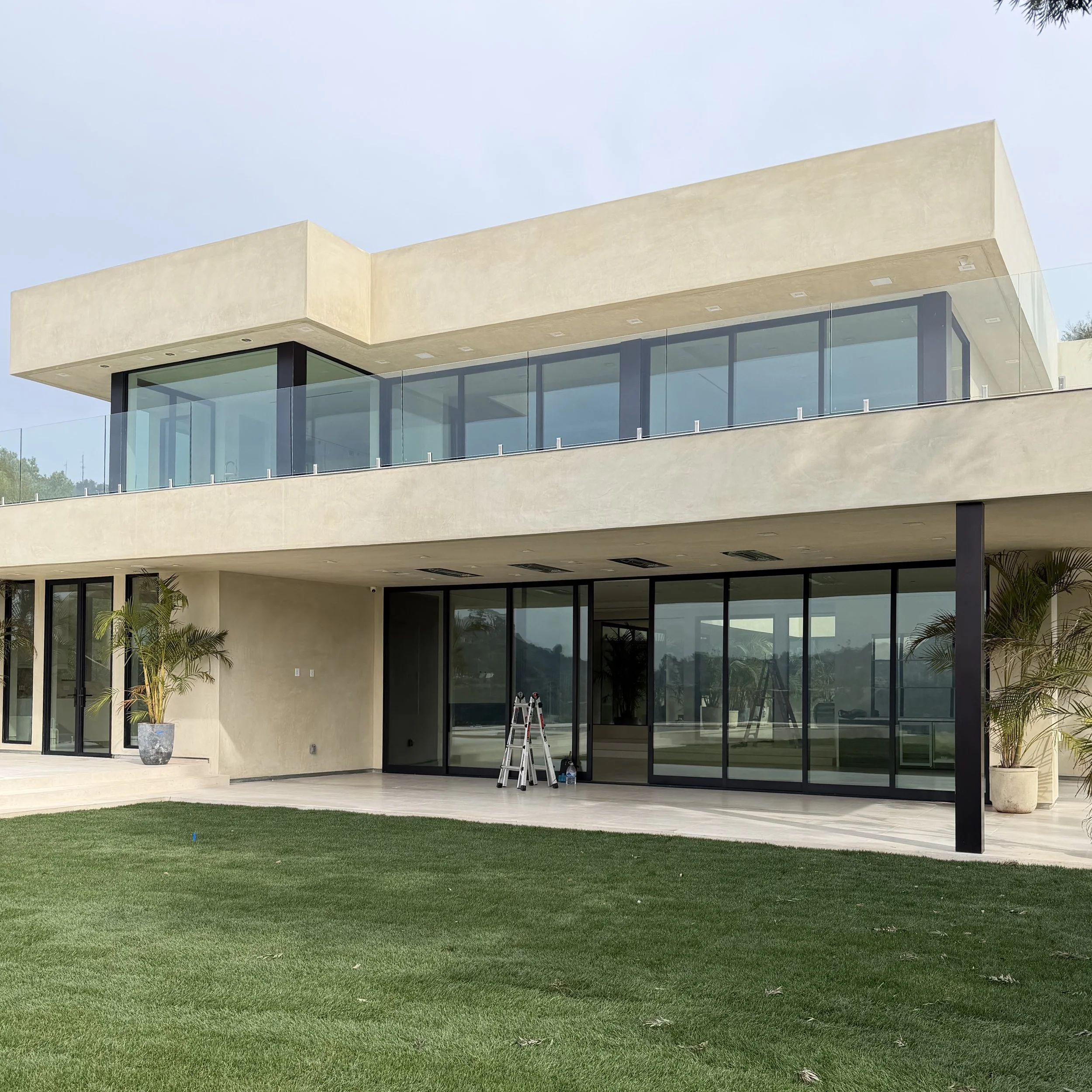 Modern multi-story house with large glass windows and sliding doors, potted plants outside, and a neatly manicured lawn in the front.