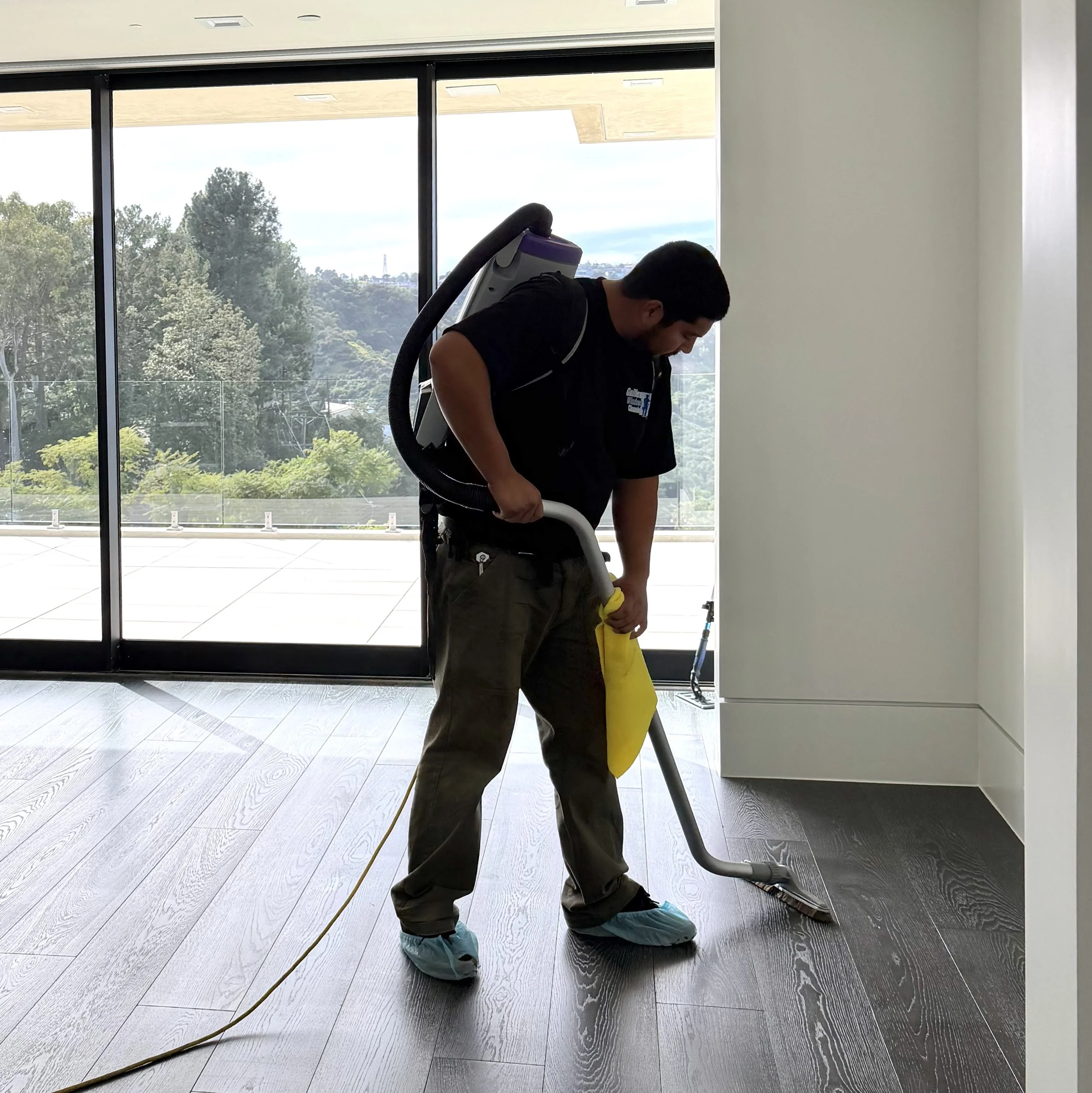 A man in protective shoe covers cleaning the dark wood floor with a floor buffer machine in a bright room with large glass sliding doors and an outdoor view of trees and hills.