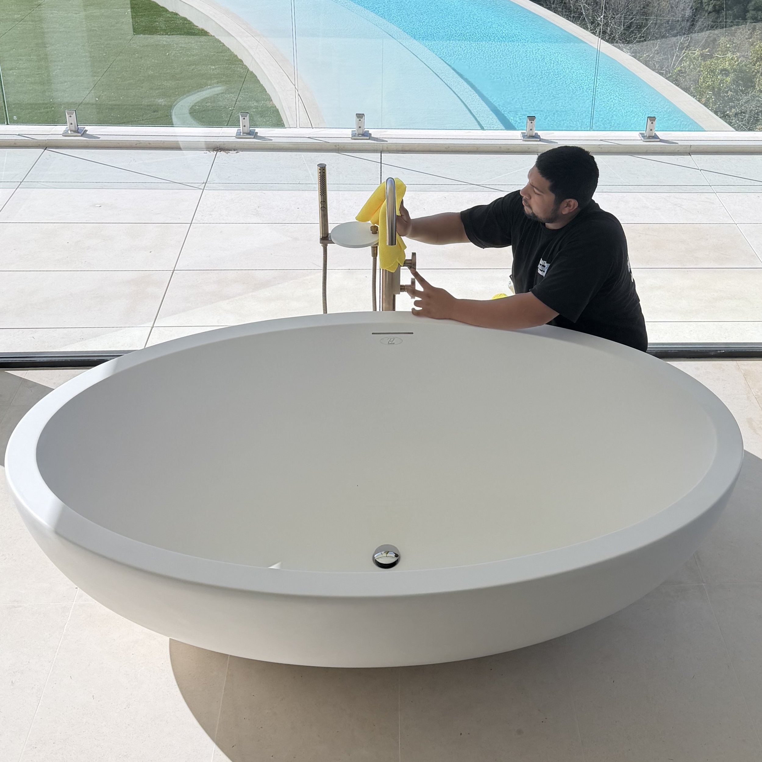 Person cleaning a large, white, round bathtub with a yellow cloth, positioned in a modern bathroom with glass walls and a view of an outdoor swimming pool.