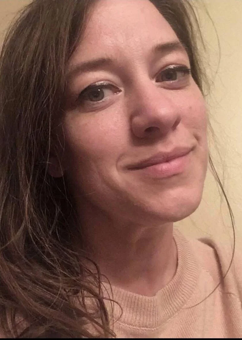 Close-up photo of a woman with brown hair and a slight smile, wearing a beige top, against a plain beige background.