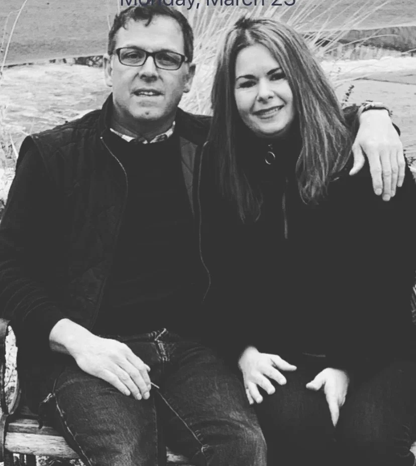 A black and white photo of a man and woman sitting on a park bench, with the woman having her arm around the man, both smiling.