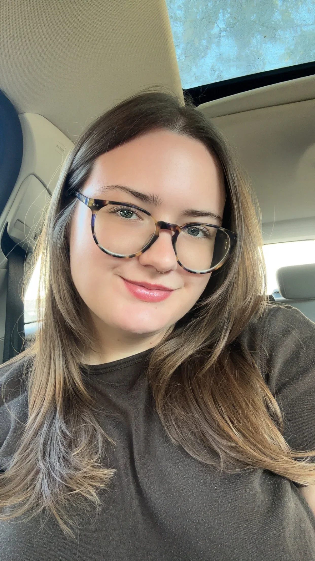 A young woman with long, wavy brown hair and glasses taking a selfie inside a car.