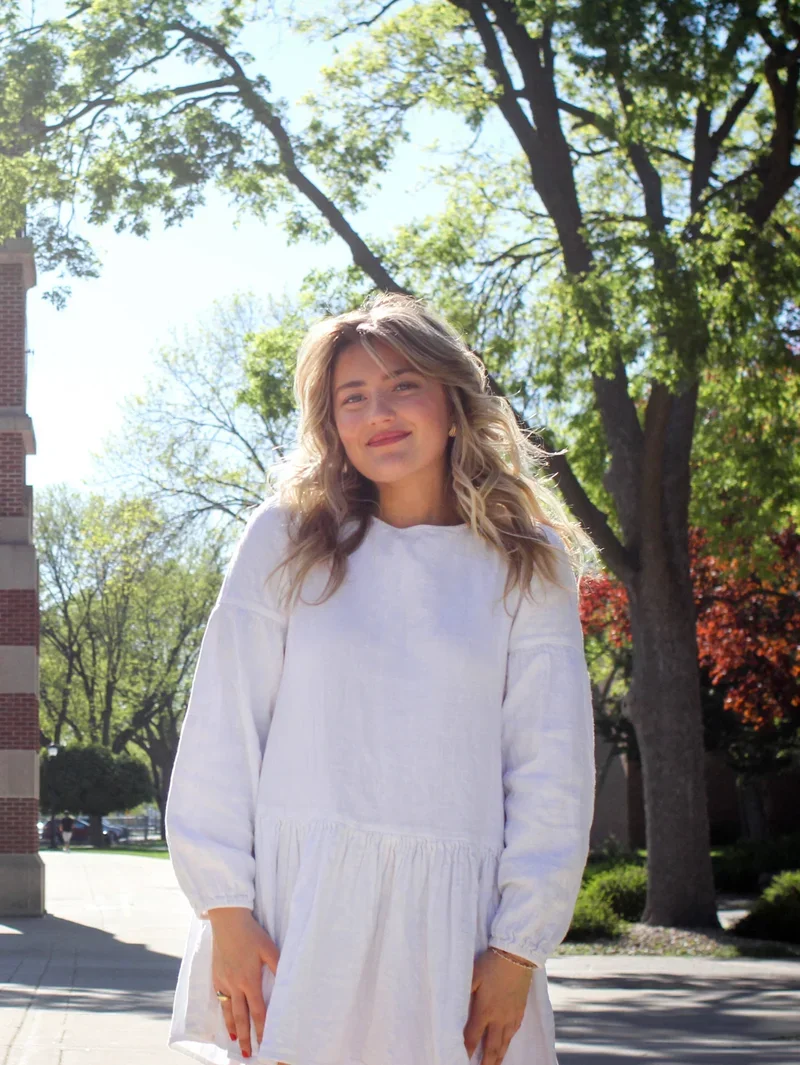A young woman with wavy blonde hair wearing a white long-sleeve dress stands outdoors on a sunny day with trees and a brick building in the background.