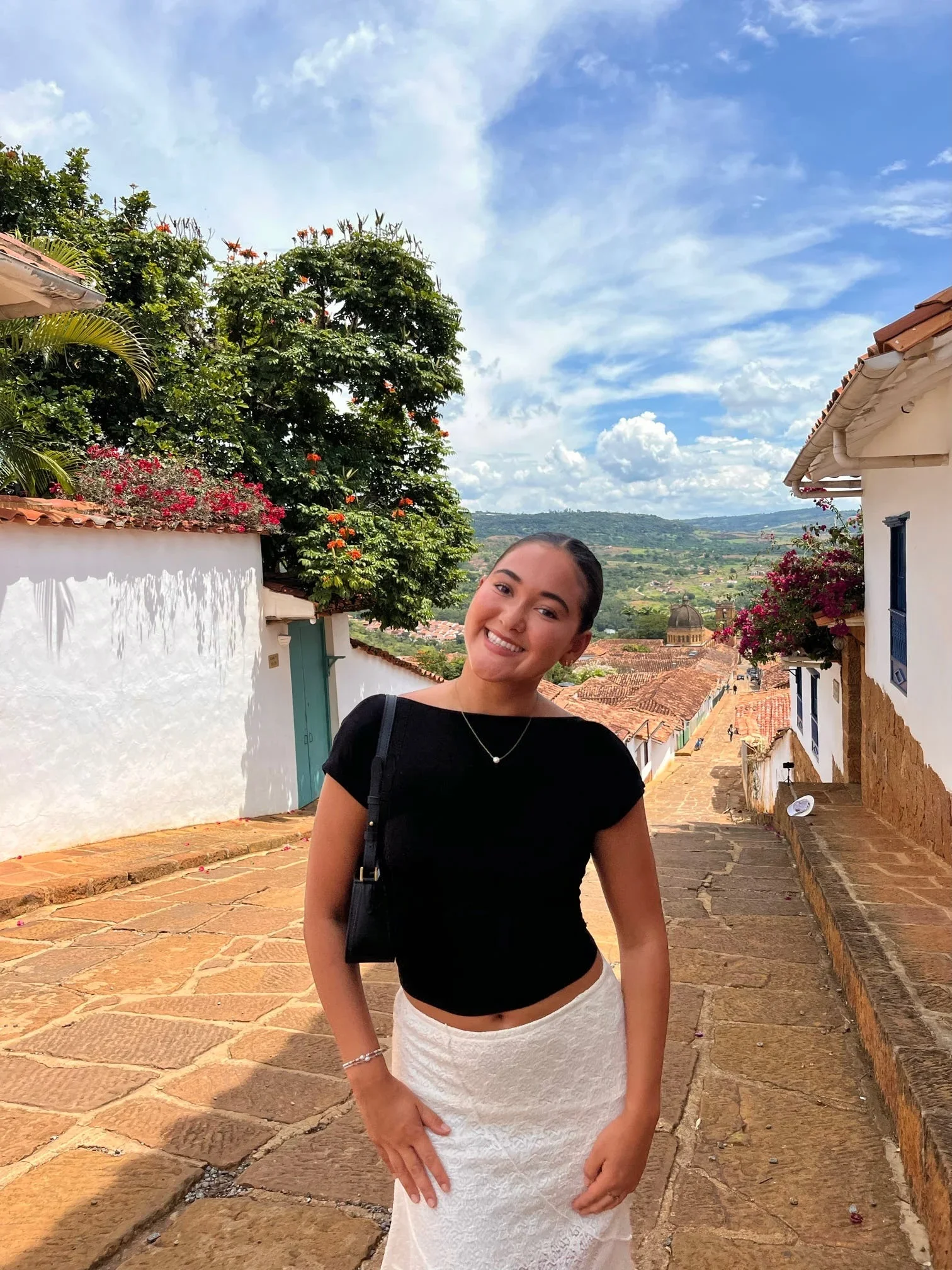 A smiling young woman with dark hair standing outdoors on a cobblestone street in a scenic village. She wears a black top and cream-colored skirt, with greenery and a blue sky with clouds in the background.