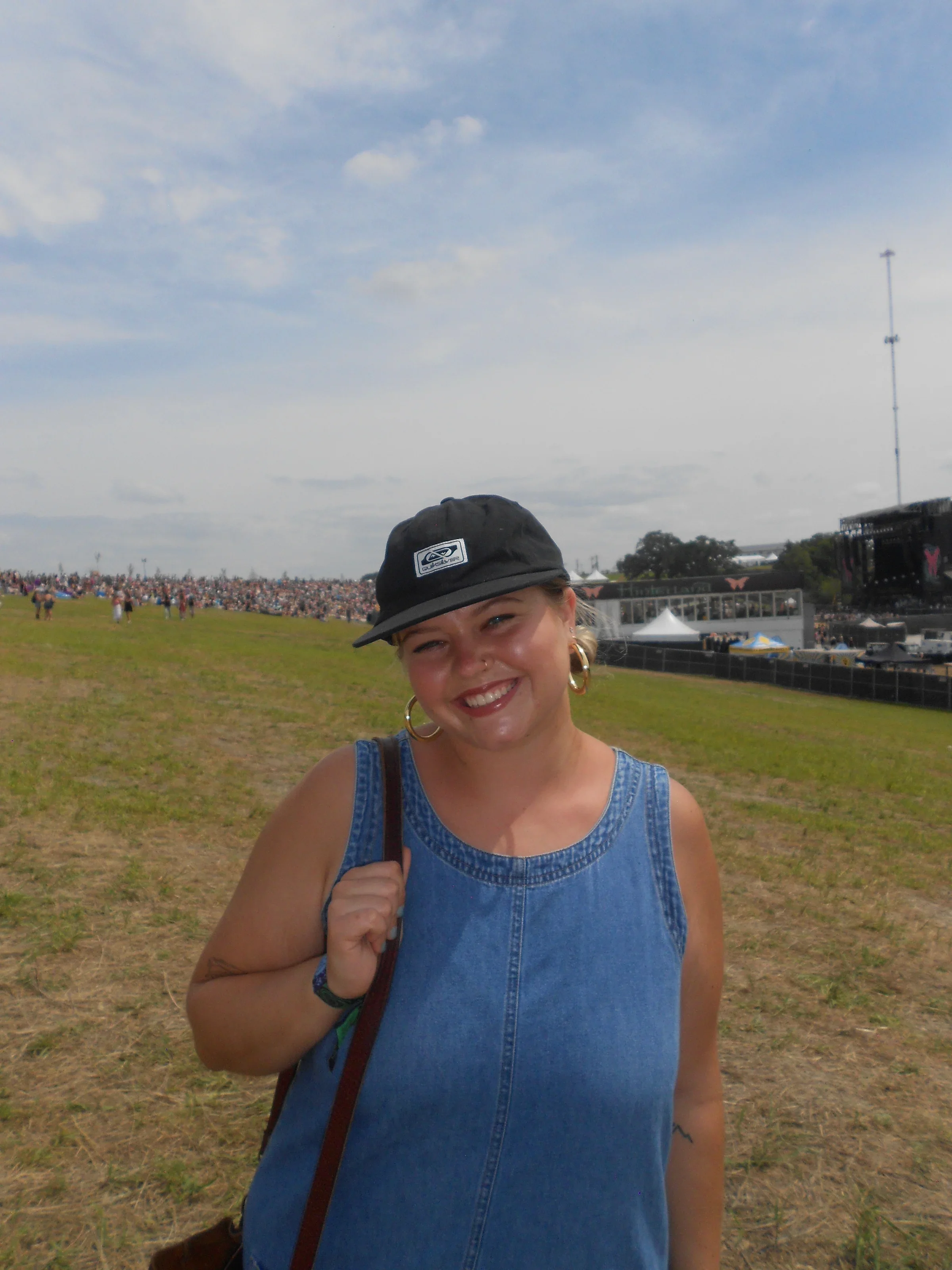 A smiling woman in a denim sleeveless top and black cap with a logo, outdoors at a large event with a stage, tents, and a crowd in the background.