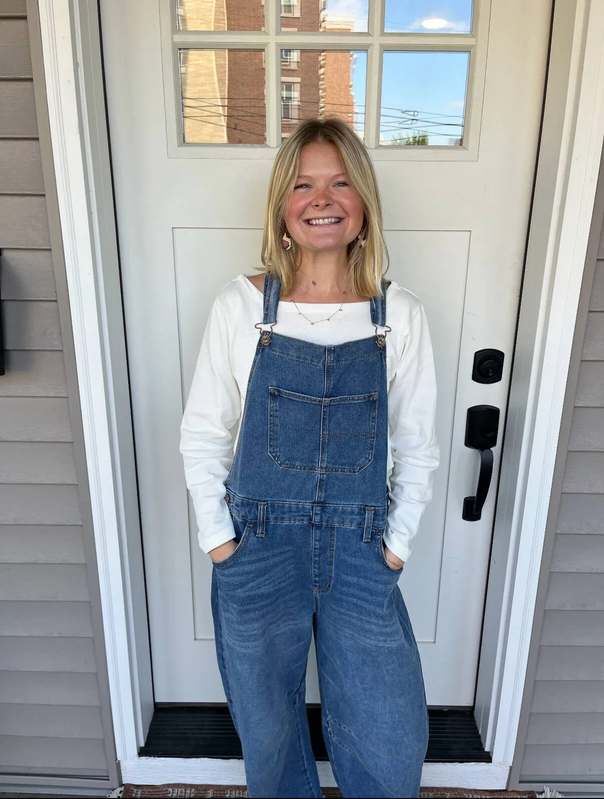 A young woman with shoulder-length blonde hair smiling and wearing a white long-sleeve shirt and denim overalls stands in front of a white door with a window, outside a house with beige siding.