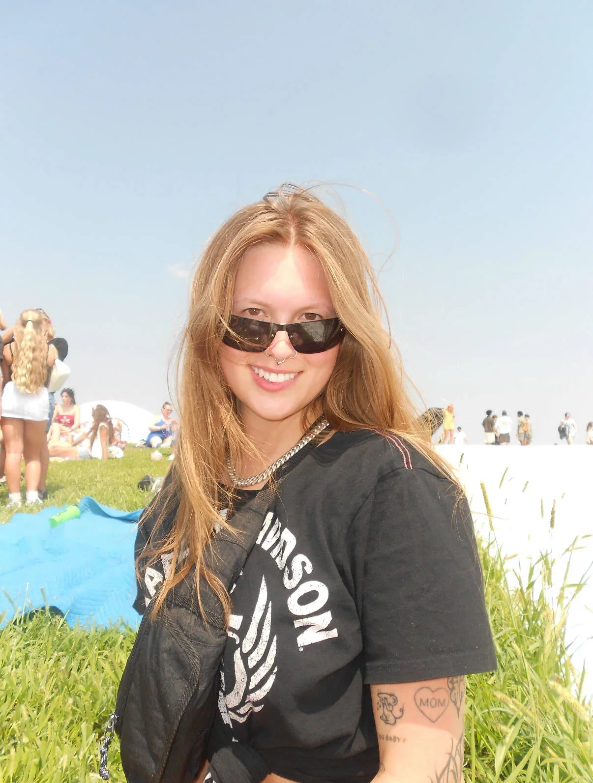 A young woman with long red hair, wearing black sunglasses, a black T-shirt with white text, and a silver chain necklace, smiling at the camera in an outdoor grassy area with people in the background and blue sky.