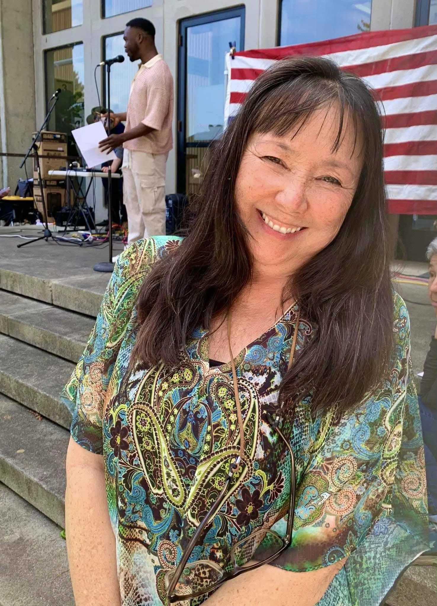 Ami (Asian American woman with long, dark hair) smiling on steps of a government building with a young African American man in background. Upside down flag behind. At a rally.