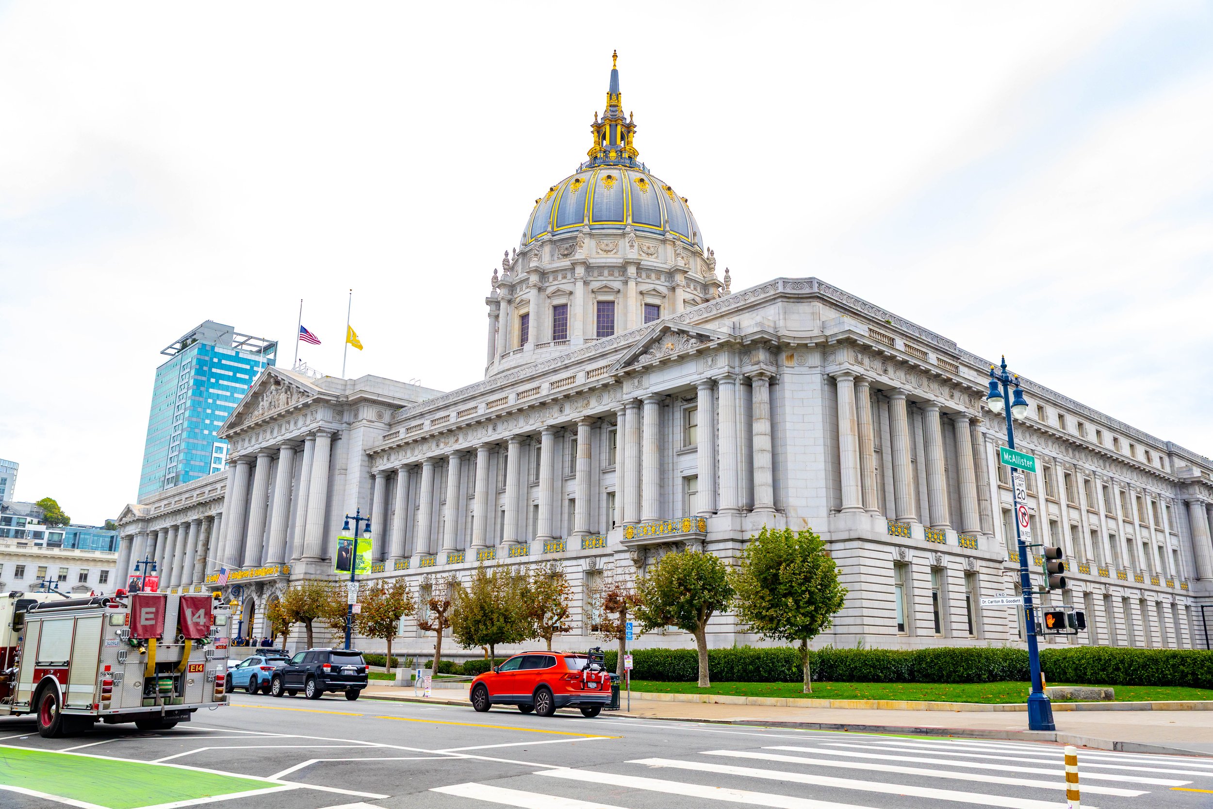 View of a large, ornate City Hall building with a golden dome, surrounded by trees and street traffic, including emergency vehicles, in San Francisco.