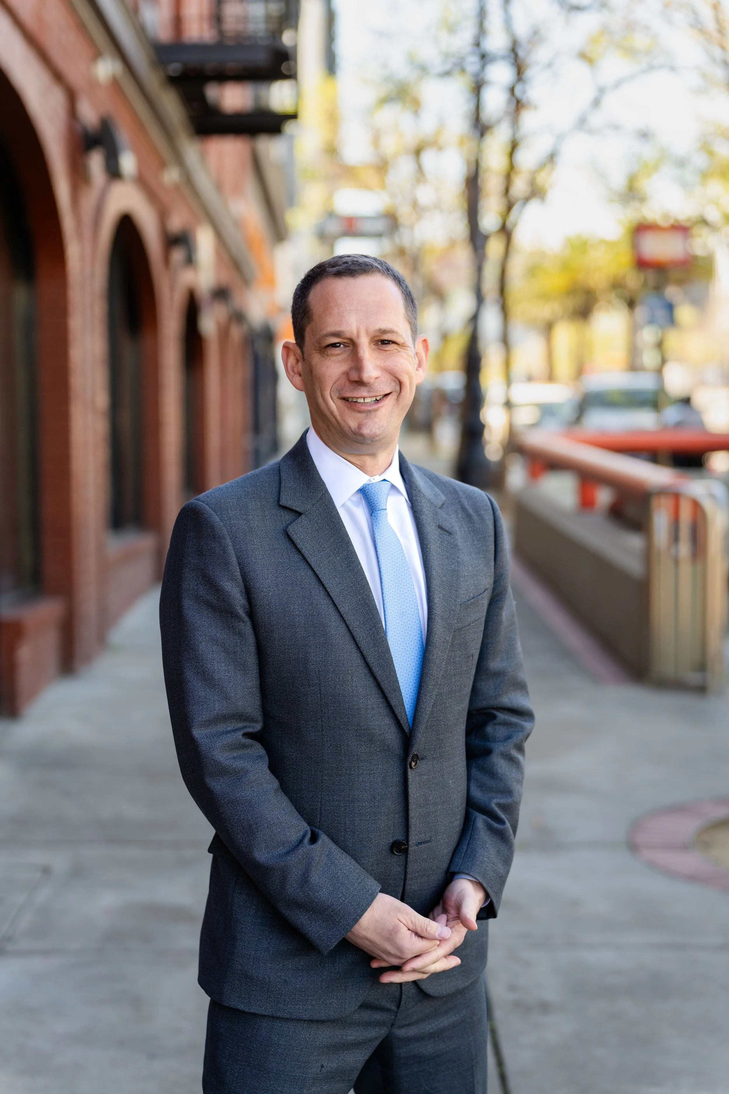 A smiling person in a gray suit and light blue tie.
