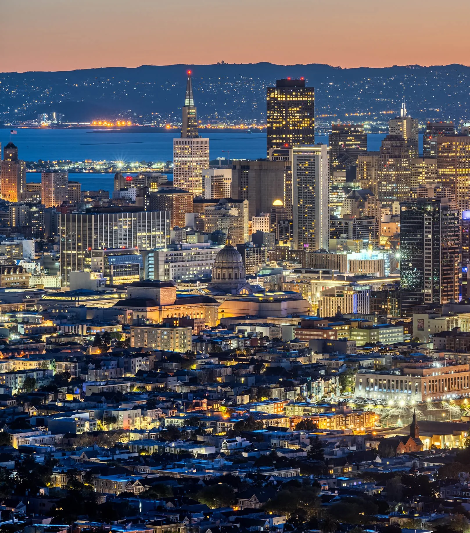 Nighttime cityscape of San Francisco with illuminated skyscrapers, waterfront, and hills in the background.