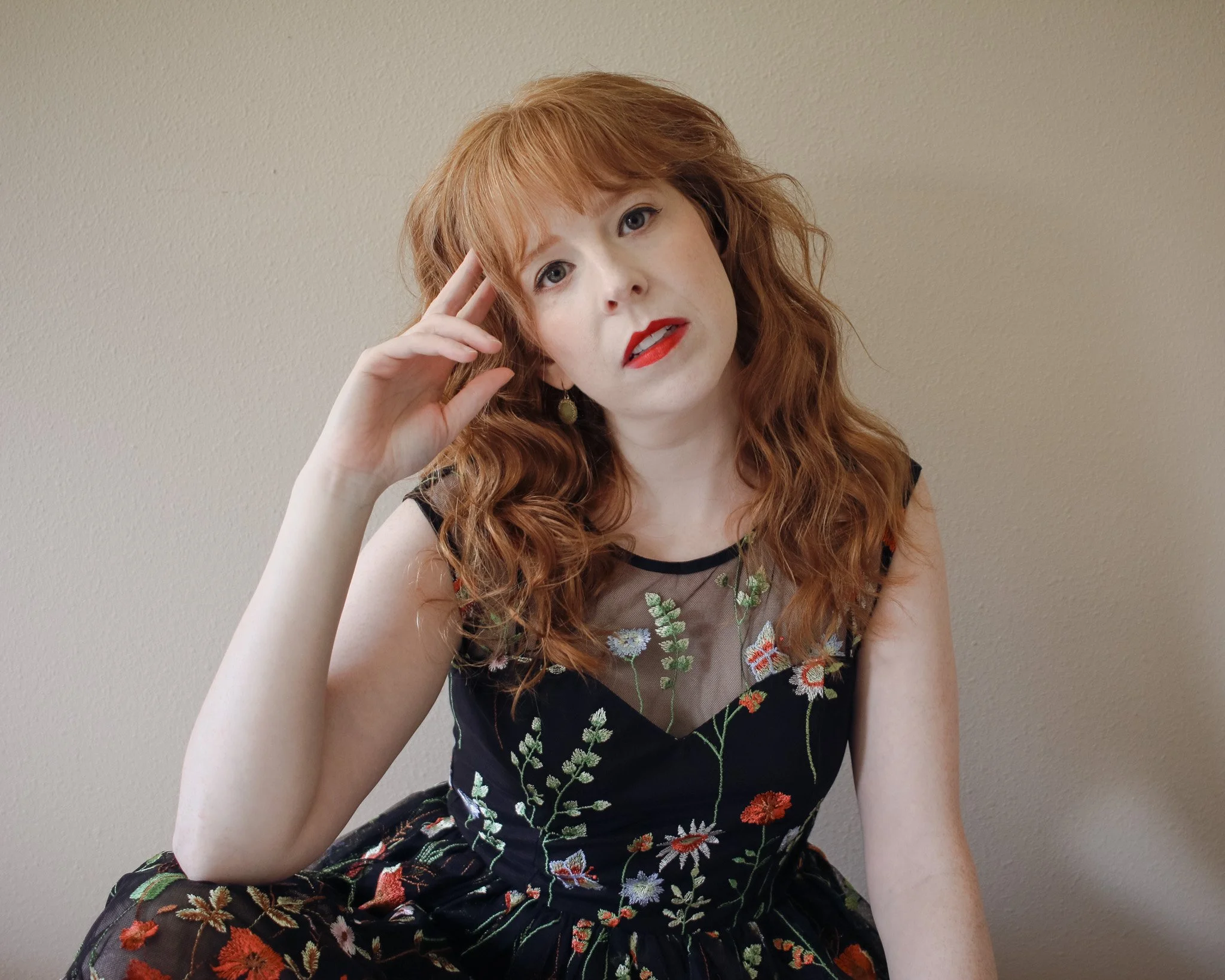 K.E. Ormsbee, fantasy author, is a 30-something white woman with curly red hair. She wears a floral dress and stares directly at the camera, head tilted to one side.