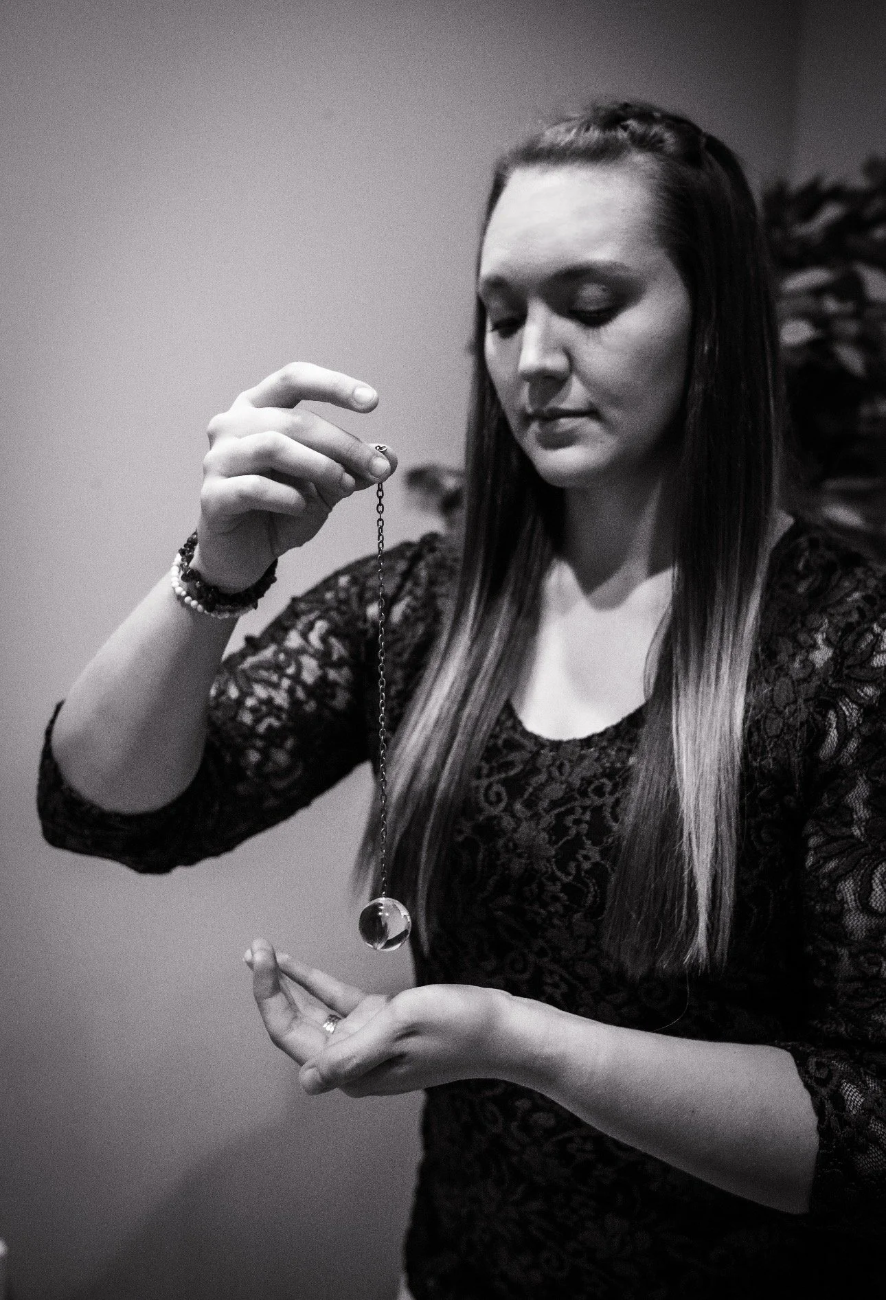 A woman with long hair, wearing a black lace dress, holding a pendulum on a chain with both hands, looking at it thoughtfully.