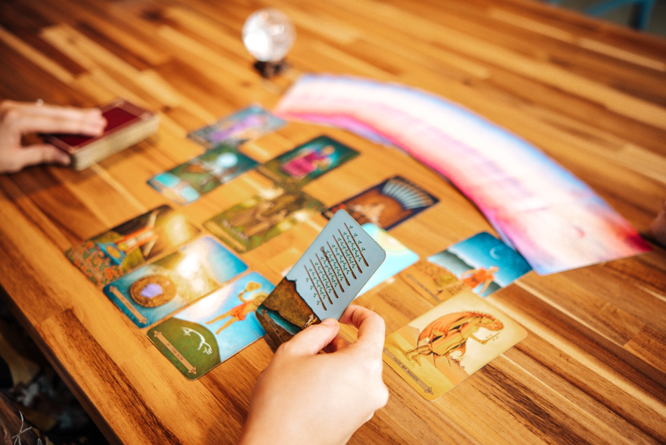 People playing a card game or tarot card reading on a wooden table with several colorful cards spread out. One person is holding a card with text on it, while another has a hand near additional cards.
