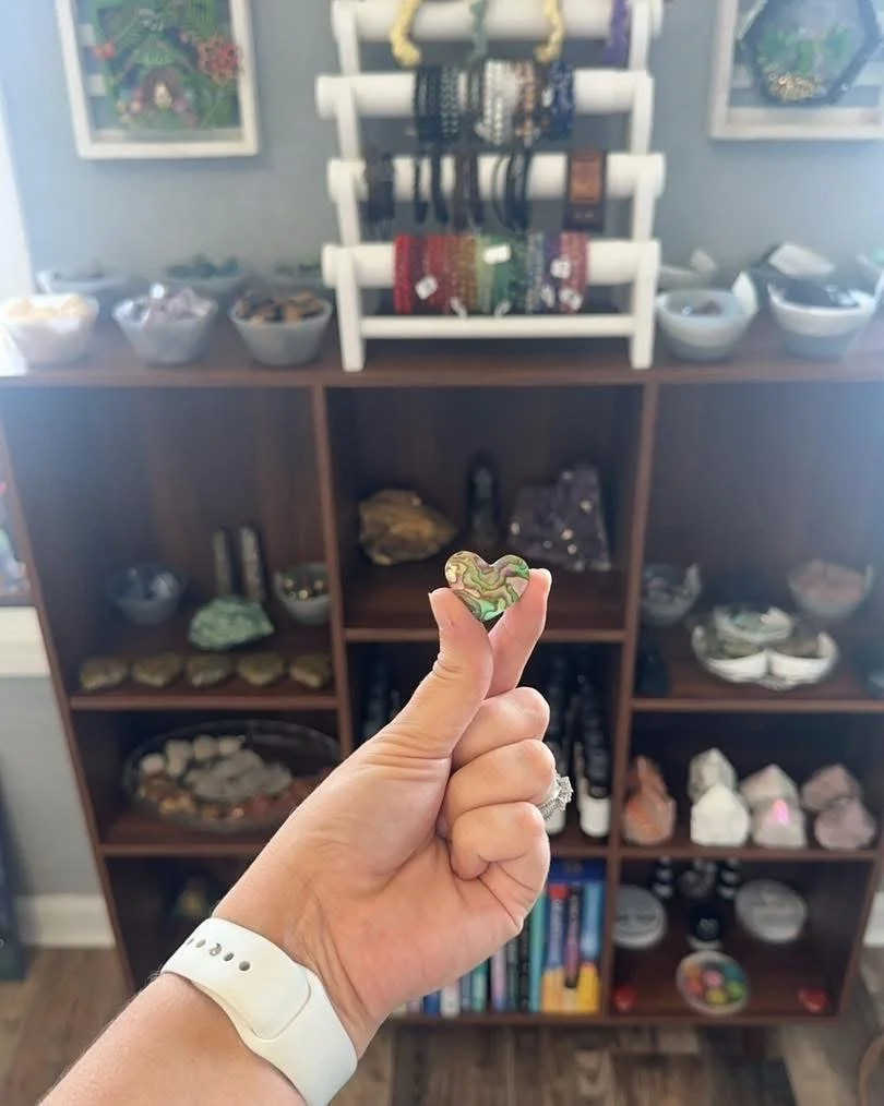 Person holding a multicolored heart-shaped object in front of a wooden display shelf with various stones, jewelry, and decorative items.