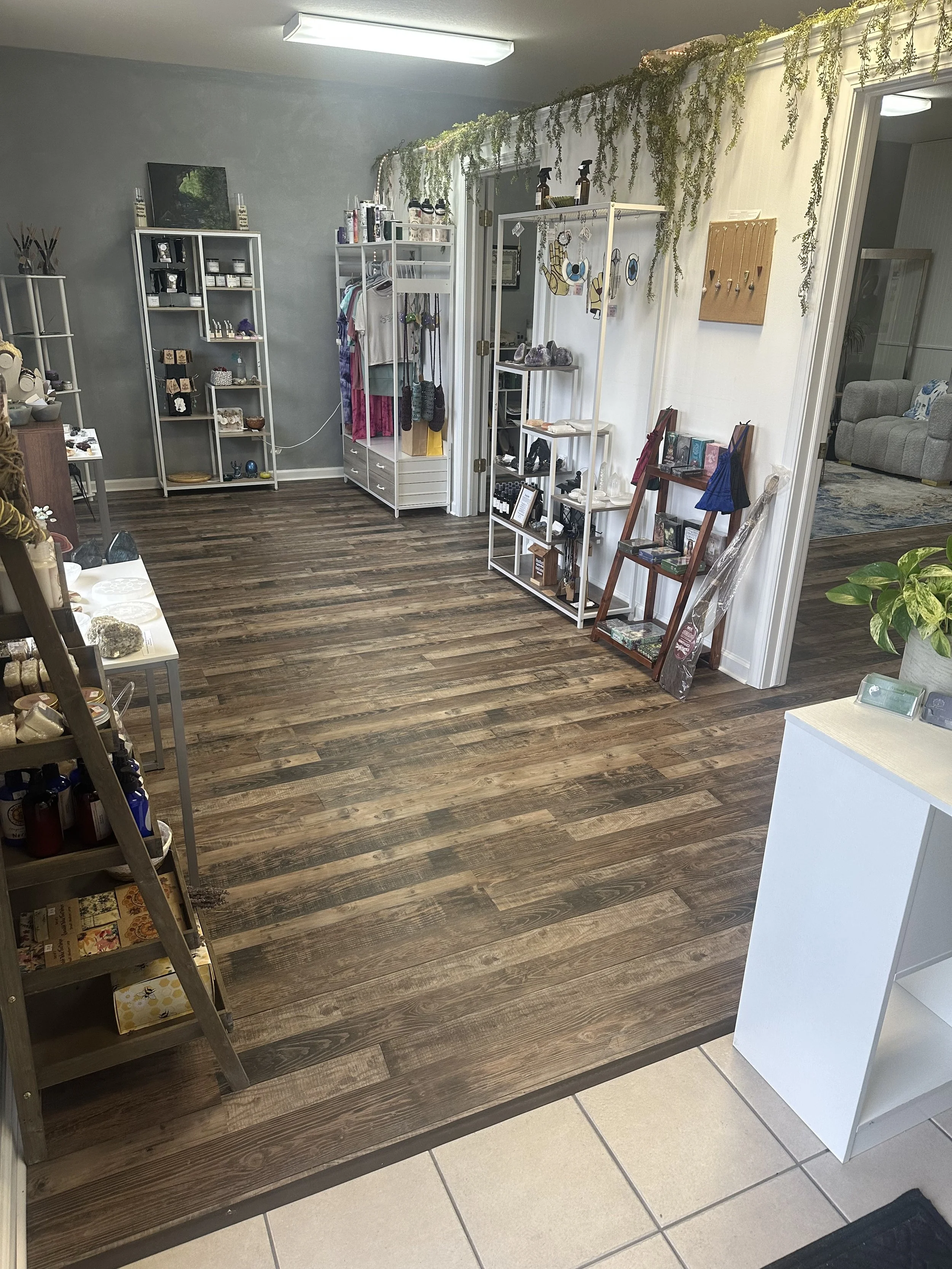 Indoor boutique or gift shop with white shelving units displaying jewelry, accessories, and souvenirs, and clothing on hangers, with wooden and tiled flooring, decorated with hanging greenery.