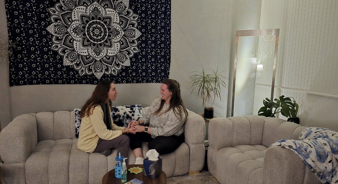 Two women sitting on a beige sofa holding hands and smiling at each other in a living room. There is a tapestry with a black and white mandala design on the wall behind them, a tall mirror, a potted plant, and a second empty beige sofa.