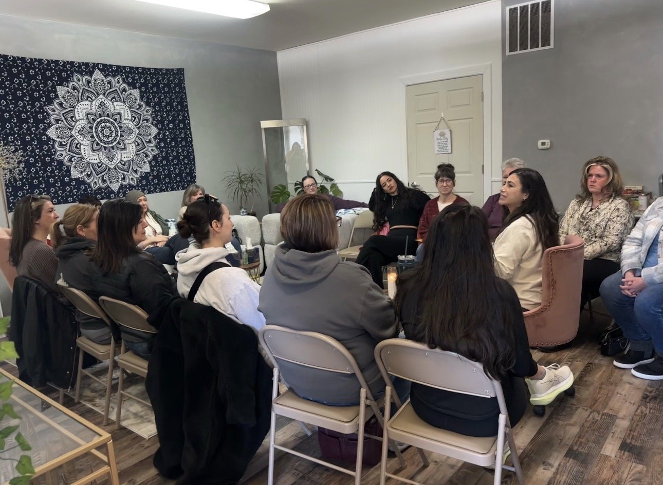A group of women gathered in a living room for a meeting or discussion, seated in a semi-circle around a coffee table.