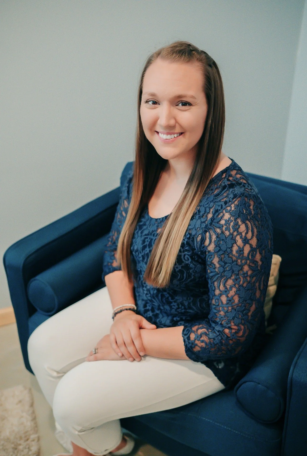 A woman with long brown hair smiling, seated on a blue sofa, wearing a dark blue lace top and white pants.