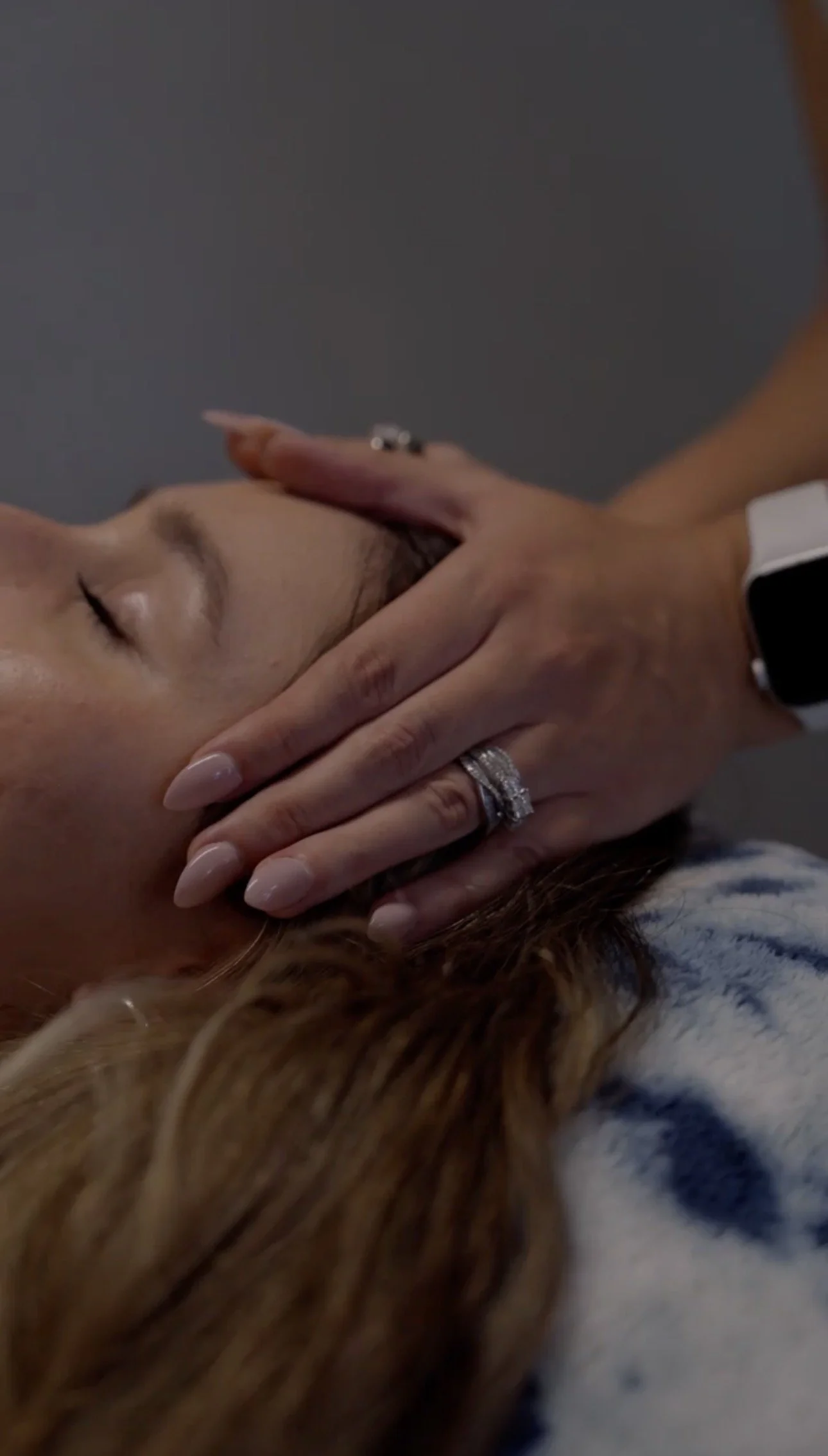 Person receiving a facial massage with their eyes closed, lying down, with long hair and wearing rings and a smartwatch.