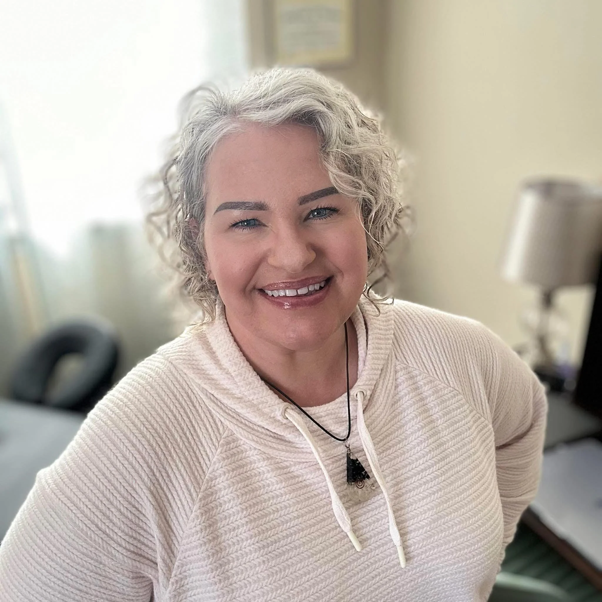 A smiling woman with curly, gray hair, wearing a cream-colored hoodie and a black necklace with a triangle pendant, standing in an office environment.