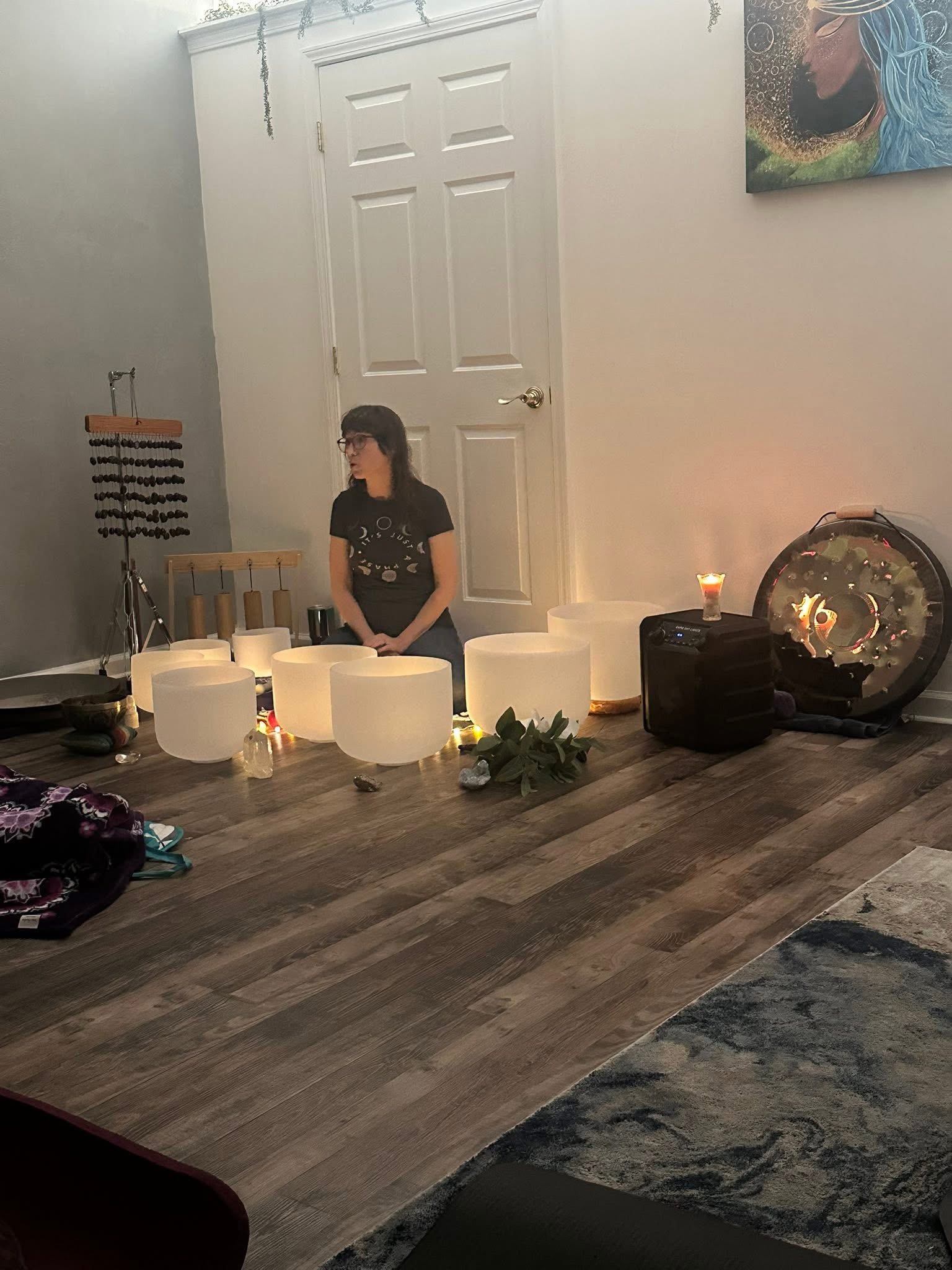 A woman with glasses sits on the floor surrounded by singing bowls, candles, and decorative stones in a room with artistic wall decor and a door in the background.