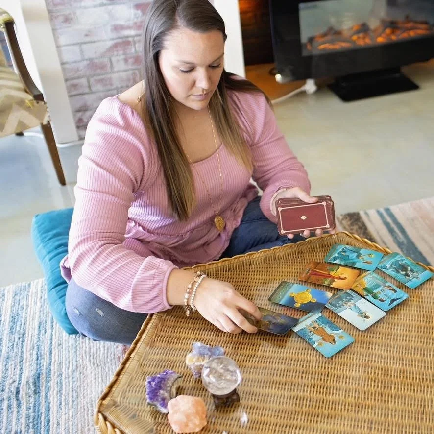 A woman in a pink top sitting on a blue cushion on the floor, reading tarot cards from a deck laid on a wicker table, with crystals in front of her and a fireplace in the background.