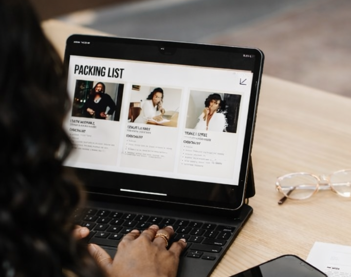 Pessoa usando um tablet com uma lista de embalagem na tela, ao lado de óculos de leitura em uma mesa de madeira.