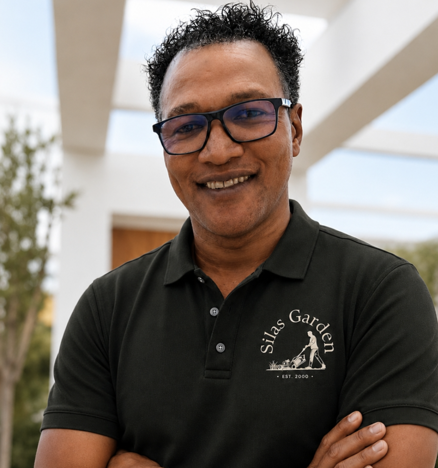 Homem sorrindo usando óculos de armação grossa e camiseta preta com logo de uma empresa de jardinagem, em ambiente ao ar livre com estrutura moderna ao fundo.