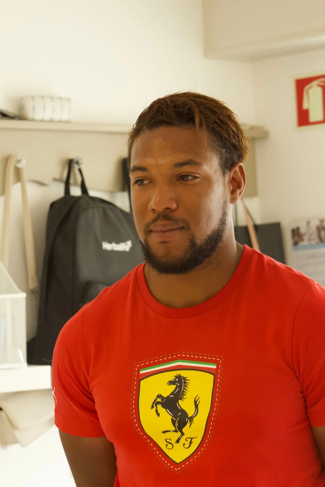 Um homem com barba, cabelo castanho escuro e pele morena, usando uma camiseta vermelha com o escudo da Ferrari em destaque. Ele está em um ambiente interno, com uma mochila preta ao fundo, em uma parede branca.