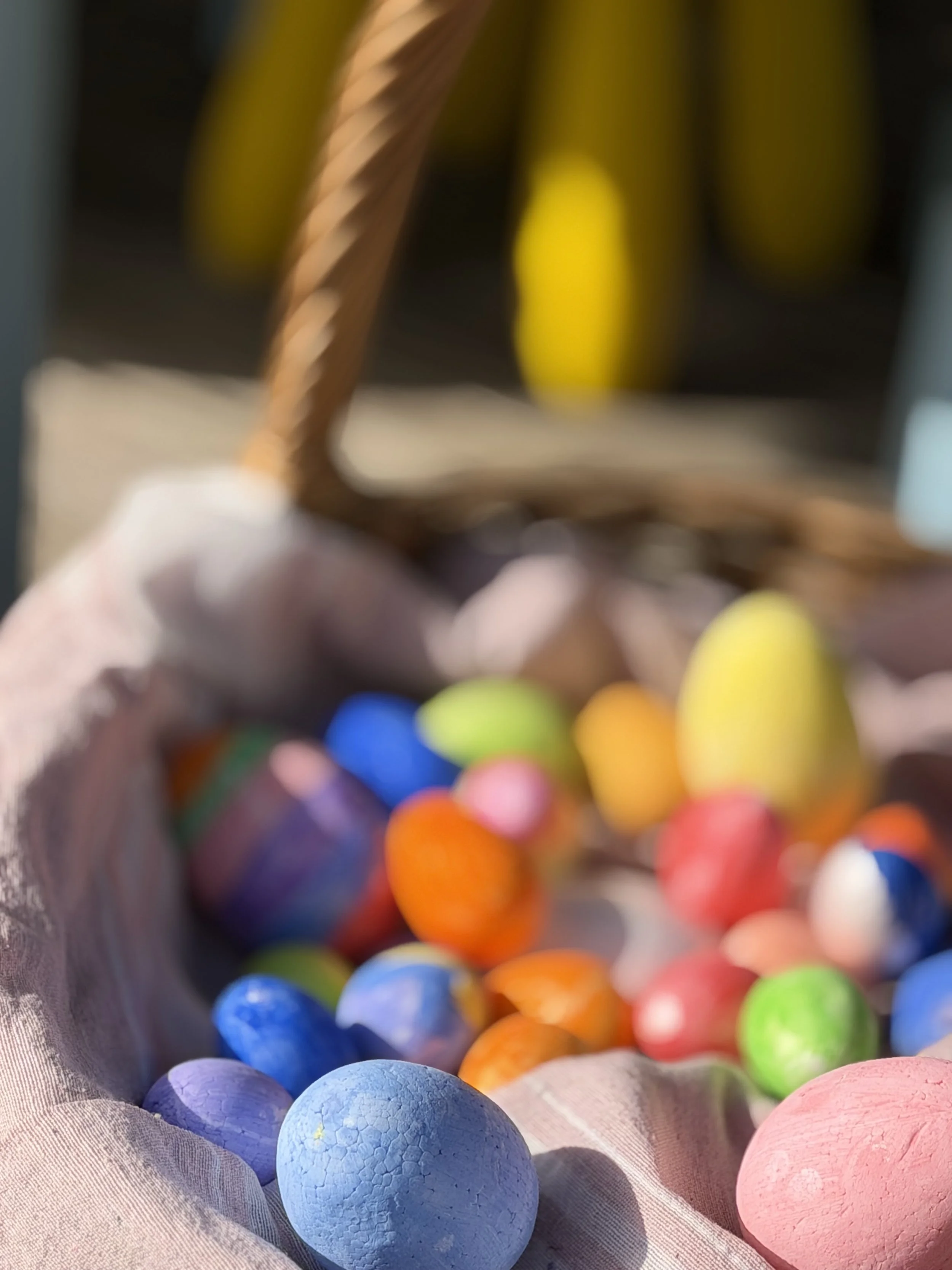 Cesta de vime com ovos de Páscoa coloridos, alguns com cores sólidas e outros com pinturas, sobre tecido de cor claro.
