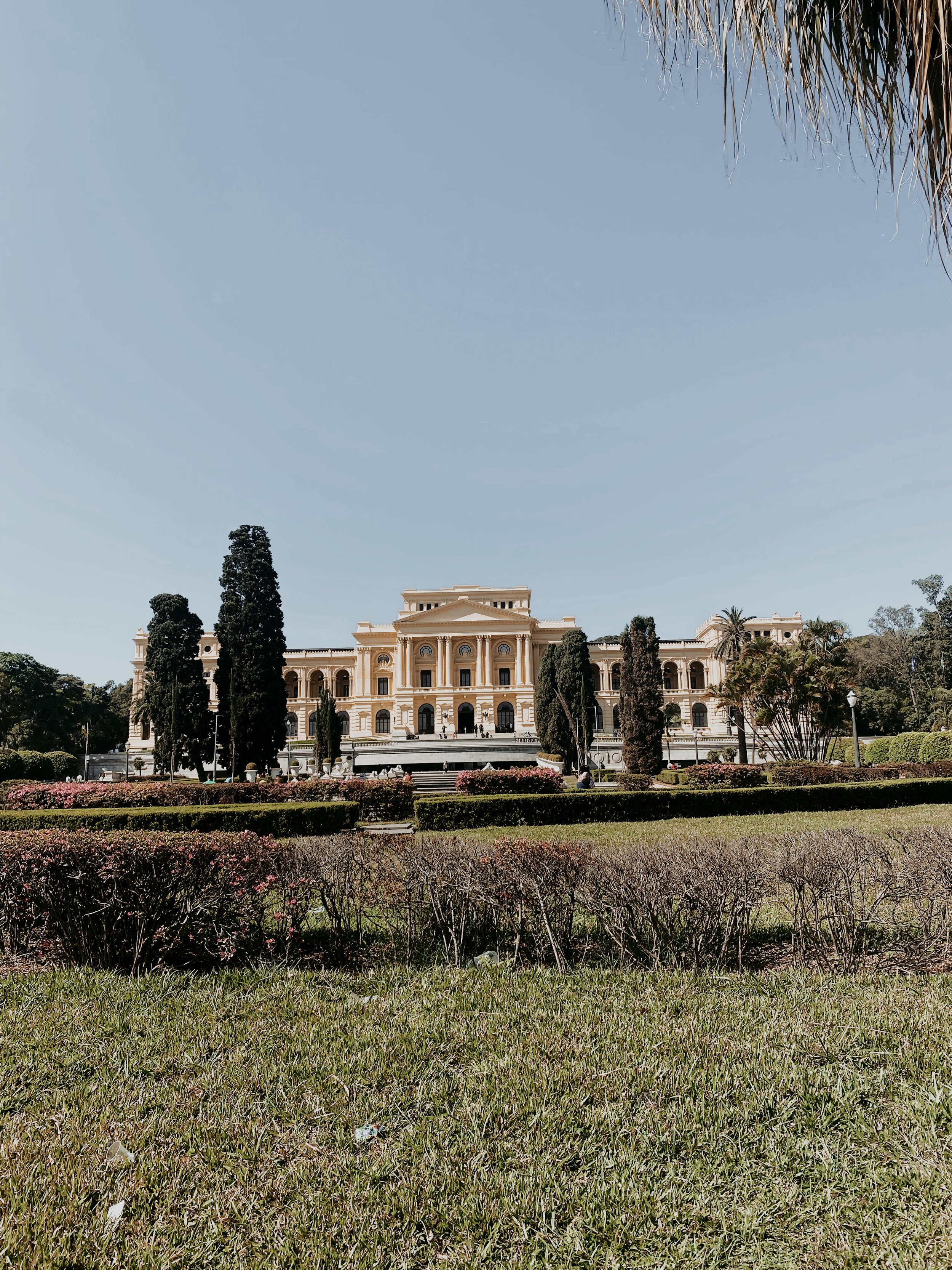 Palácio de estilo neoclássico rodeado de jardins com arbustos, árvores altas e gramado sob céu claro.