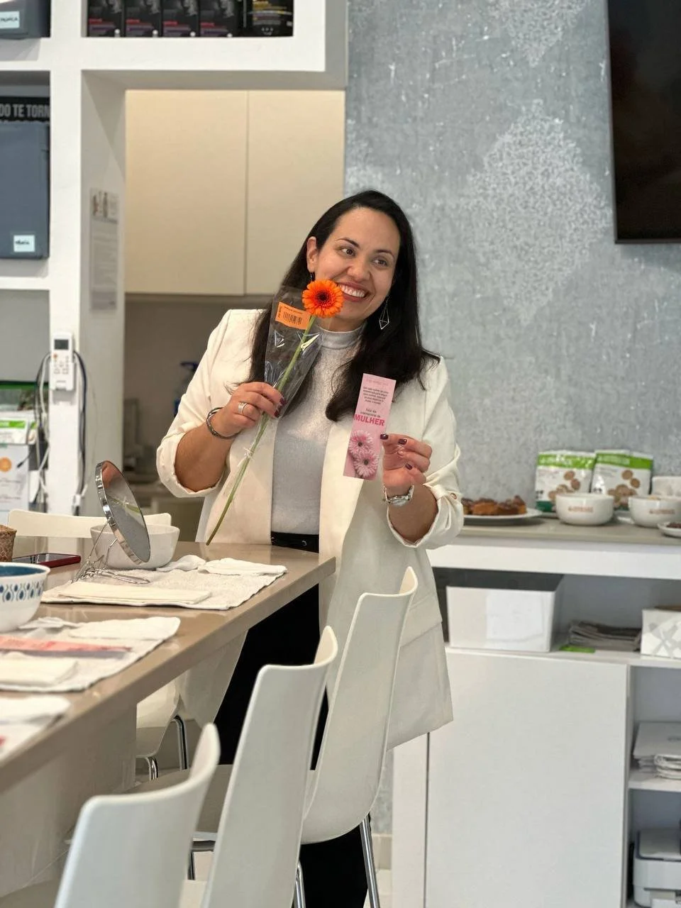 Mulher sorridente segurando uma flor de cor laranja, com uma credencial na mão, em um ambiente de cozinha ou sala de jantar, com cadeiras brancas e utensílios na mesa.