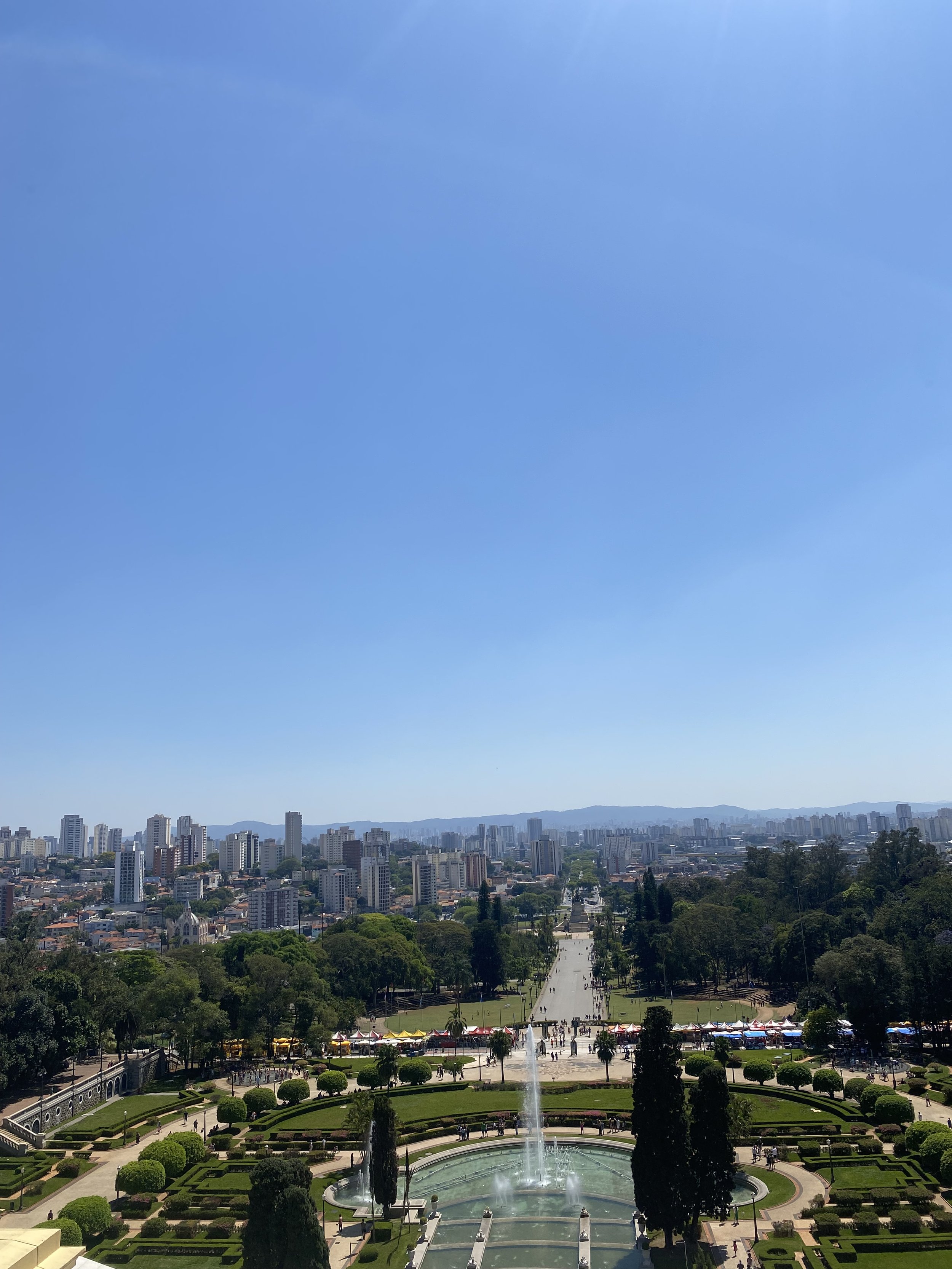 Vista de um parque com fontes, árvores bem cuidadas e uma ampla avenida no centro, com várias pessoas caminhando, e uma cidade com prédios altos ao fundo sob um céu claro