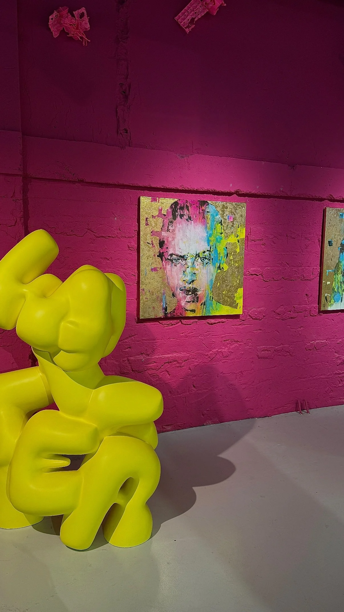 Escultura amarela de uma mão gigante formando um dedo do meio, em frente a parede rosa com quadros de arte contemporânea, incluindo uma pintura de rosto feminino