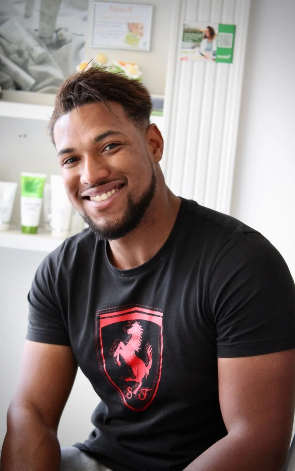 Homem sorridente com cabelo castanho claro, barba bem feita, usando uma camiseta preta com logotipo de cavalo vermelho e letras 'S' e 'F'.
