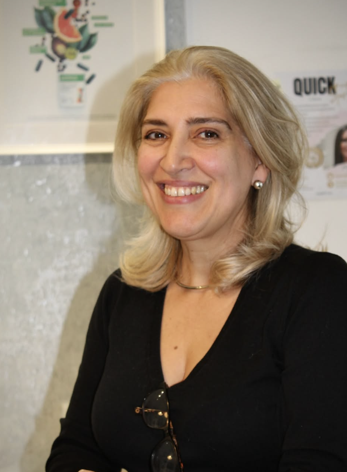 Mulher sorridente com cabelo loiro, usando blusa preta, brincos de pérola, e um colar, posando dentro de um ambiente com parede de textura clara ao fundo, com telas e avisos.