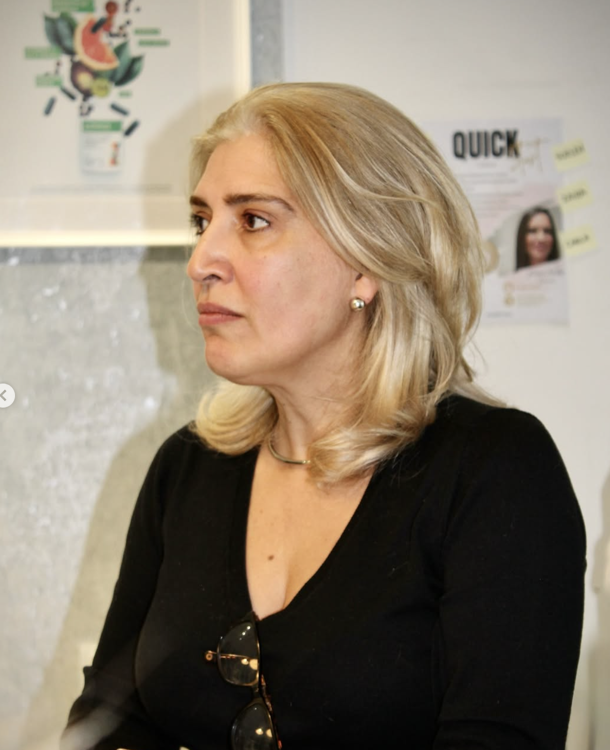 Mulher com cabelo loiro, vestindo uma blusa preta, usando brincos de pérola, em ambiente interno com parede de fundo com cartazes e anúncios.