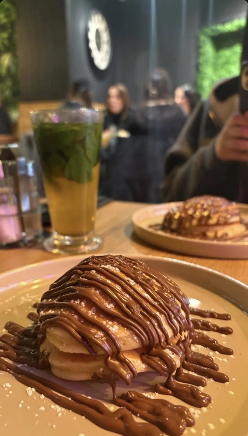 Prato de sobremesa com panquecas cobertas com calda de chocolate e chantilly, ao fundo uma bebida verdejar, possivelmente com hortelã, em um restaurante com pessoas ao fundo.