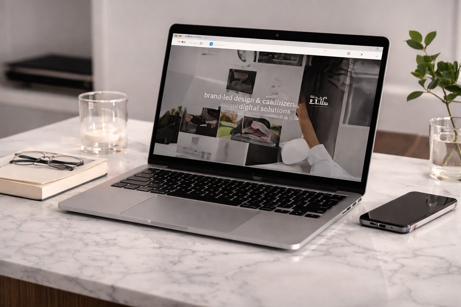 Laptop no centro da mesa mostrando uma página de design digital, ao lado estão um caderno com óculos, uma vela acesa, um smartphone e uma planta em um vaso de vidro.