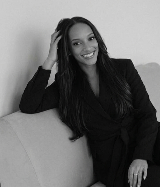 Mulher sorrindo, com cabelo longo e escuro, sentada no sofá, usando um blazer preto, em um ambiente interno.